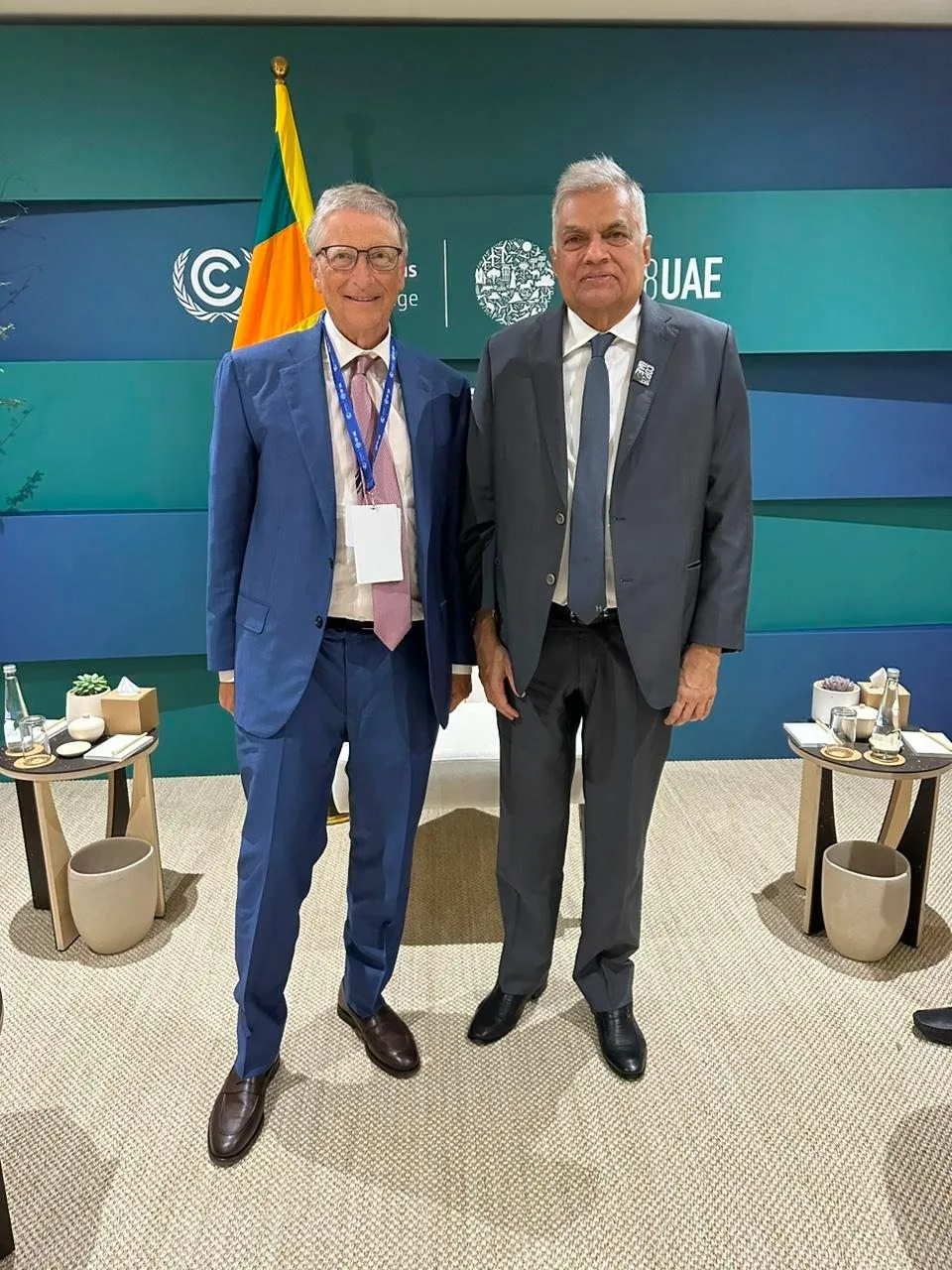 Bill Gates and Ranil Wickremesinghe are posing together in a formal setting, likely at a conference or diplomatic event. Both individuals are dressed in business attire, with Bill Gates wearing a blue suit, a light-colored shirt, and a pink tie, complemented by brown shoes. Ranil Wickremesinghe is in a dark grey suit with a white shirt and a dark tie, paired with black shoes. The setting appears to be indoors, characterized by a modern, corporate design.
Behind them, a backdrop features a logo and the word "UAE," suggesting the event could be related to the United Arab Emirates. A flag is prominently displayed, adding a diplomatic tone to the scene. The room is furnished with small tables holding water bottles and decorative plants, indicating a formal meeting environment. The mood seems cordial, with both individuals smiling slightly, reflecting a positive interaction. The lighting is bright, enhancing the professional atmosphere of the occasion.