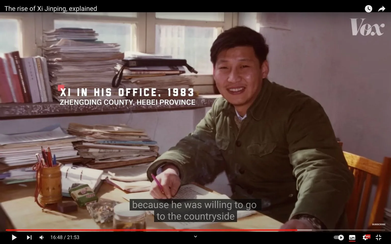 Young Xi Jinping is depicted in this video frame, inside an office setting in 1983, located in Zhengding County, Hebei Province. The scene shows him seated at a desk, exuding a confident and approachable demeanor as he smiles towards the camera. He is dressed in a green military-style jacket, which is typical of the era and setting, suggesting a formal yet practical attire. The office is cluttered with stacks of papers and books, indicating a busy and engaged work environment. In front of him, there is a collection of pens in a holder and several other office supplies, reflecting an active workspace. The lighting is warm and natural, coming from a window behind him, creating a comfortable and inviting atmosphere. The video is part of a broader narrative by Vox, titled "The rise of Xi Jinping, explained," which explores the historical and political context of Xi Jinping’s early career. The frame is accompanied by subtitles that read, "because he was willing to go to the countryside," hinting at a narrative of dedication and willingness to work in rural areas.
