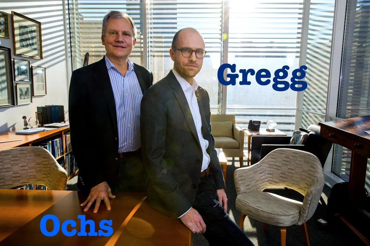 The image features two men, identified by the hint as "Sulzberger father and son," standing in a modern office environment. The man on the left, labeled "Ochs," wears a dark blazer over a blue striped shirt, exuding a professional demeanor with a gentle smile. To his right, the man labeled "Gregg" also wears a dark blazer, paired with a white shirt and glasses, conveying a more serious expression. Both are standing beside a wooden table, with sunlight filtering through the blinds in the background, casting lines of shadow across the room.
The office is well-lit, with large windows offering a glimpse of the cityscape outside. The furnishings include a comfortable-looking chair, a desk with various objects, and a bookshelf filled with books, giving the space a professional yet personal feel. The overall mood is formal yet approachable, highlighting a connection between the two men, who are dressed similarly and positioned closely, suggesting a familial and business relationship. The presence of their names in bold blue text adds emphasis to their identities.