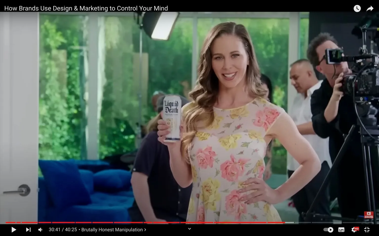Cherie Deville is prominently featured in this video frame from YouTube, titled "How Brands Use Design & Marketing to Control Your Mind." She is posing with a can labeled "Liquid Death," smiling confidently at the camera. Cherie is wearing a floral dress with a pattern of pink and yellow roses, exuding a vibrant and cheerful vibe. Her hair is styled in loose waves, and she stands with one hand on her hip, embodying a poised and professional demeanor suitable for a commercial setting.
In the background, a filming setup is visible, including a cameraman operating professional video equipment. The setting provides a glimpse into a typical behind-the-scenes environment during the production of a commercial. The lighting is bright, highlighting Cherie and the product she is holding. The room's large windows reveal a green, leafy view outside, adding a natural element to the scene. The overall atmosphere suggests a lively and dynamic shoot, emphasizing the focus on marketing and branding techniques.
