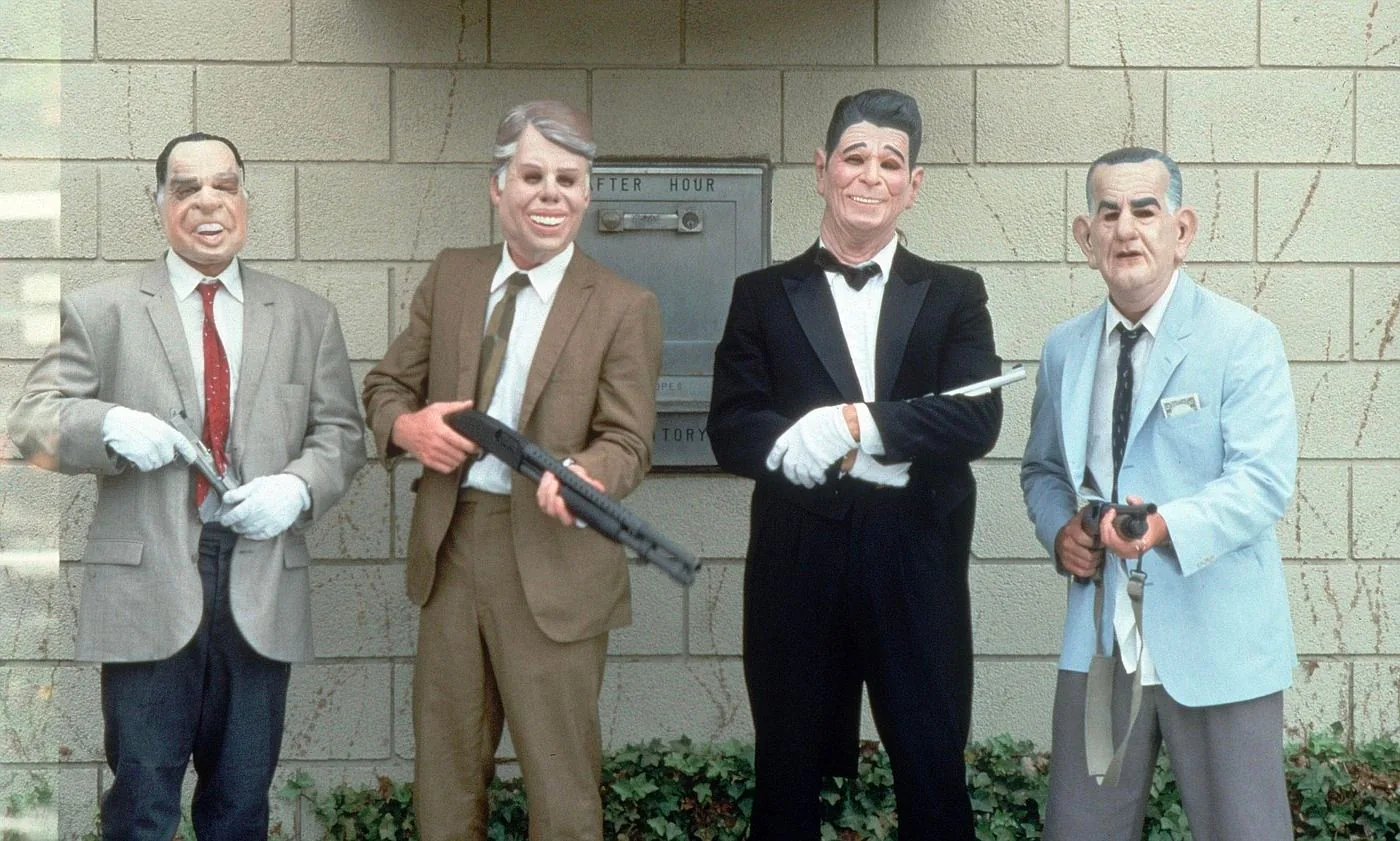 The image depicts a scene from the film "Point Break," showing four individuals wearing masks resembling past U.S. presidents. They are standing in front of a brick wall with foliage at the bottom. Each person is dressed in formal or semi-formal attire, with the third individual from the left sporting a black tuxedo. They are holding various weapons, adding to the heist theme. The masks are caricature-like, adding a humorous yet menacing touch to the scene. The lighting is bright and even, suggesting the setting is outdoors during the day. There is a small mailbox or dropbox behind them labeled "AFTER HOUR," contributing to the setting's context. The overall mood is tense yet slightly comedic due to the exaggerated expressions on the masks.