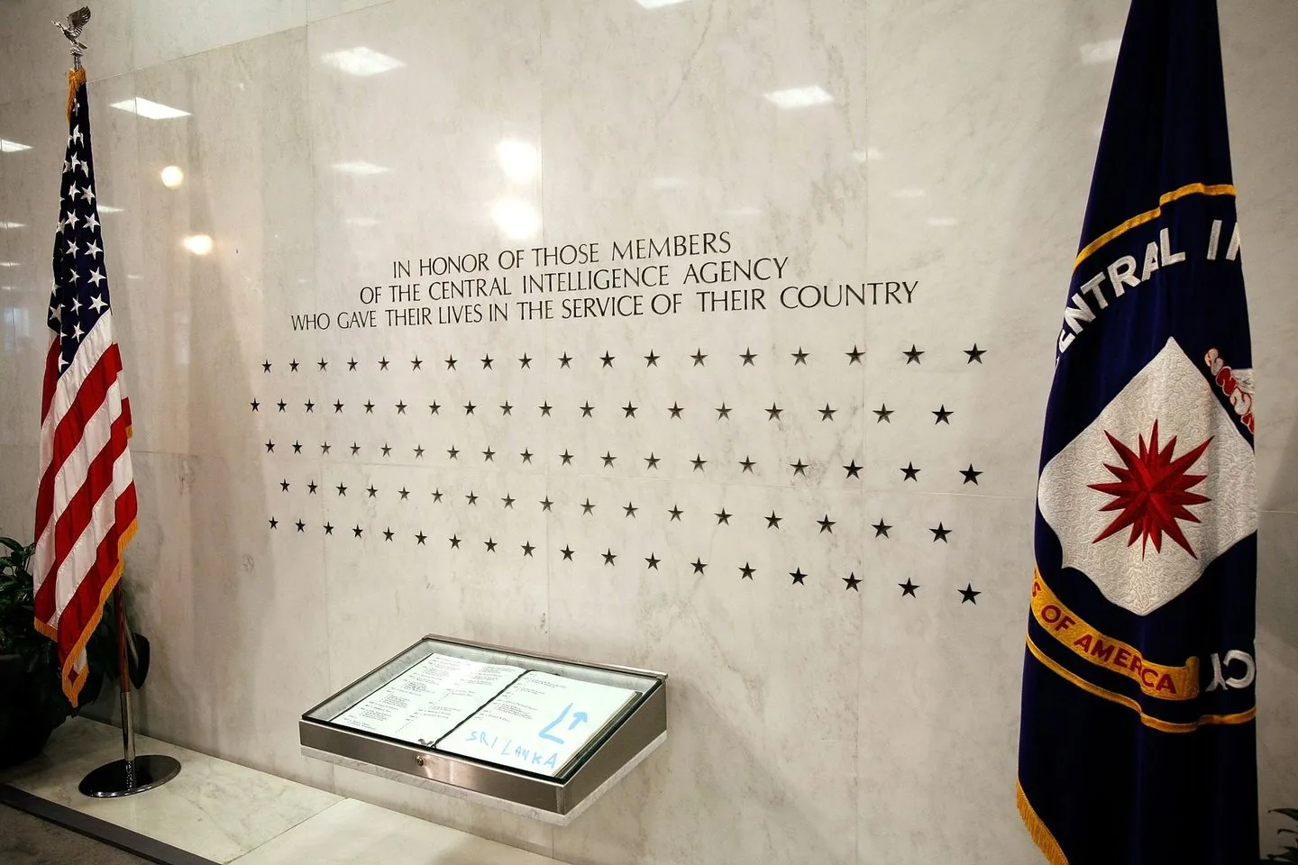 The image depicts a solemn memorial wall at the Central Intelligence Agency headquarters. The wall is made of polished marble and features several stars, each representing an individual who gave their life in service. Above the stars, there is an inscription honoring these members of the CIA. The text reads, "IN HONOR OF THOSE MEMBERS OF THE CENTRAL INTELLIGENCE AGENCY WHO GAVE THEIR LIVES IN THE SERVICE OF THEIR COUNTRY."
On the left side of the image is an American flag, standing tall on a silver pole with an eagle finial. To the right, the flag of the Central Intelligence Agency is partially visible, featuring the agency's emblem—a red compass rose set against a white background. Below the inscription, there is a glass case displaying a book, possibly containing the names of those honored by the stars. The environment is brightly lit, reflecting off the smooth, clean surfaces of the wall and floor, creating a respectful and dignified atmosphere befitting a memorial.