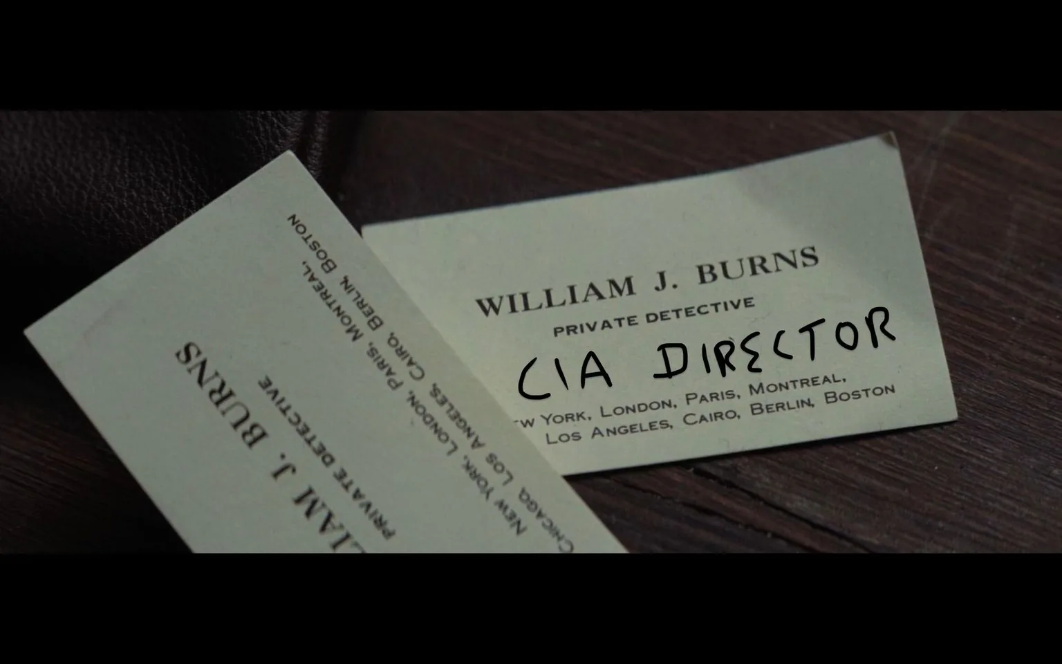 In this image, there is a close-up of a business card on a wooden surface. The card belongs to "William J. Burns," identified as a "Private Detective," with several cities listed below: New York, London, Paris, Montreal, Los Angeles, Cairo, Berlin, Boston. Notably, the text "CIA DIRECTOR" is handwritten over the printed details, altering the professional identity indicated on the card. The card's design is simple, with a light background and dark, serif text, suggesting a formal and professional aesthetic.
The card is partially overlapping another business card with similar design elements, hinting at a setting where multiple cards might be considered or exchanged, perhaps in an investigative or diplomatic context. The lighting is soft, emphasizing the text and the tactile quality of the paper, creating an atmosphere of intrigue or mystery. The handwritten addition introduces an element of humor or irony, playing with the idea of identity and authority.