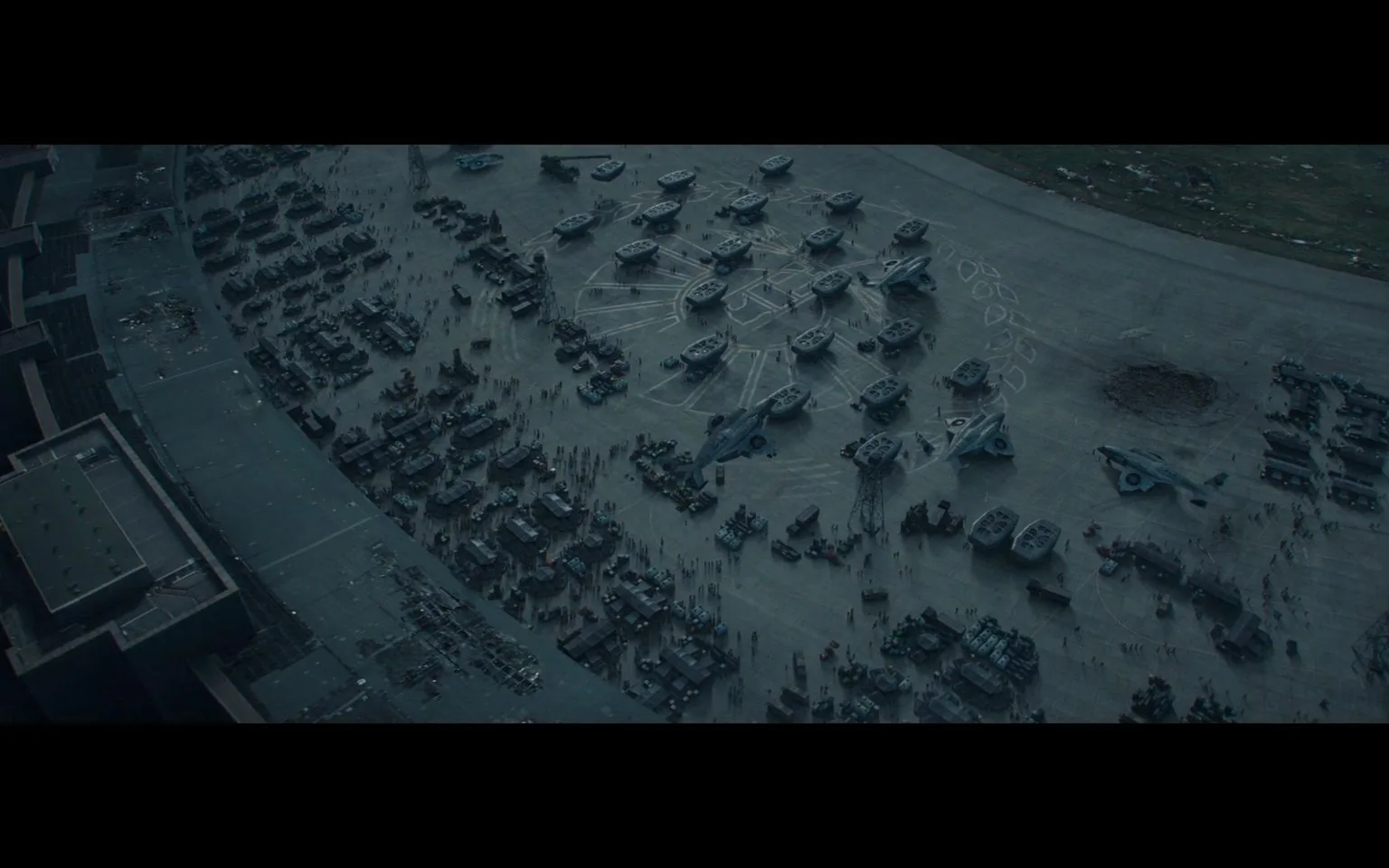 This image features an aerial view of a vast military base. The scene is dominated by numerous military vehicles and aircraft parked in orderly rows on a large concrete surface. The aircraft resemble futuristic or fictional models, with distinctive designs, possibly suggesting a setting from a science fiction or military-themed film. Surrounding the aircraft are various other military vehicles, including trucks and armored units.
The organization of the vehicles suggests a well-structured military operation or preparation for deployment. There are small clusters of people scattered throughout the area, indicating activity and movement, although individual details are not discernible from the distance at which the scene is captured. The lighting is subdued, with a muted color palette that conveys a sense of seriousness and tension. In the background, there are hints of additional structures and open land, reinforcing the notion of a sprawling, strategic military installation. The overall mood is one of preparation and readiness.