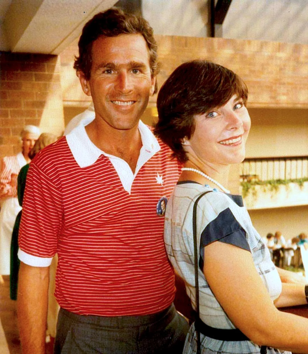 ```
Laura Bush is seen in an indoor setting, attending what appears to be a casual event. She is wearing a dress with a pattern that includes shades of blue and white, and she accessorizes with a pearl necklace. Her expression is friendly and she is slightly turned toward the camera. Standing beside her is a man in a red and white striped polo shirt with short sleeves, which includes a small emblem on the chest. The background shows a warmly lit room with other attendees, some of whom are dressed in similar casual attire, suggesting a relaxed social gathering. The atmosphere is bright, with natural light streaming in, and the environment appears to be a public venue, possibly a hall or event space. The overall mood is convivial, with the attendees seeming to enjoy their time.