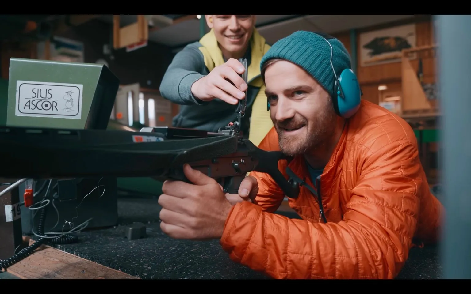 This image shows an indoor shooting range setting where two individuals are engaged in a shooting activity. The person in the foreground is lying prone, holding a rifle and aiming with focus. He's wearing a bright orange jacket and a teal beanie, complemented by blue ear protection, indicating attention and safety in the noisy environment. The expression on his face suggests concentration and enjoyment. Another individual stands beside him, assisting by adjusting something on the rifle with a small tool, smiling in a supportive manner.
The scene is well-lit, with a mix of natural and artificial lighting, providing a warm and inviting atmosphere. In the background, the interior appears to be lined with wooden panels, typical of a sports or training facility. A visible green device with the label "SIUS ASCOR" is placed prominently, indicating a system used for electronic scoring or target management. Wires and equipment are scattered on a table, emphasizing the technical and precise nature of the activity.
