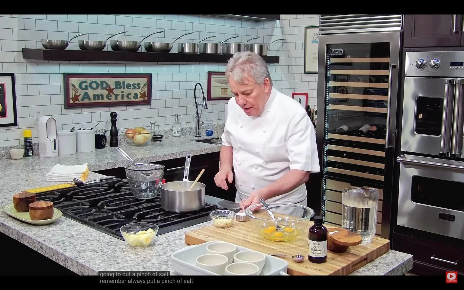 ```
SECTION 2: Rich prose
In this image, a chef is actively engaged in a cooking demonstration within a modern kitchen setting. The chef, wearing a white double-breasted chef's coat, is focused on the task at hand, preparing ingredients with precision. At the forefront is a granite countertop cluttered with various cooking essentials: a saucepan on the stove, a bowl of eggs, a container of butter, and a bottle labeled "100% Pure Tahitian Vanilla." These elements suggest a baking or dessert preparation.
The kitchen backdrop features a stainless steel wine refrigerator, prominently displaying the brand "Viking," and a tiled wall with a decorative sign reading "GOD Bless America." A series of metal mixing bowls are neatly lined up on a wall shelf, adding to the culinary ambiance. The lighting is bright, emphasizing the cleanliness and organization of the space. The chef's expression and posture convey concentration, underscoring the instructional nature of the scene. Subtitles at the bottom highlight a focus on seasoning, with a reminder to always add a pinch of salt.