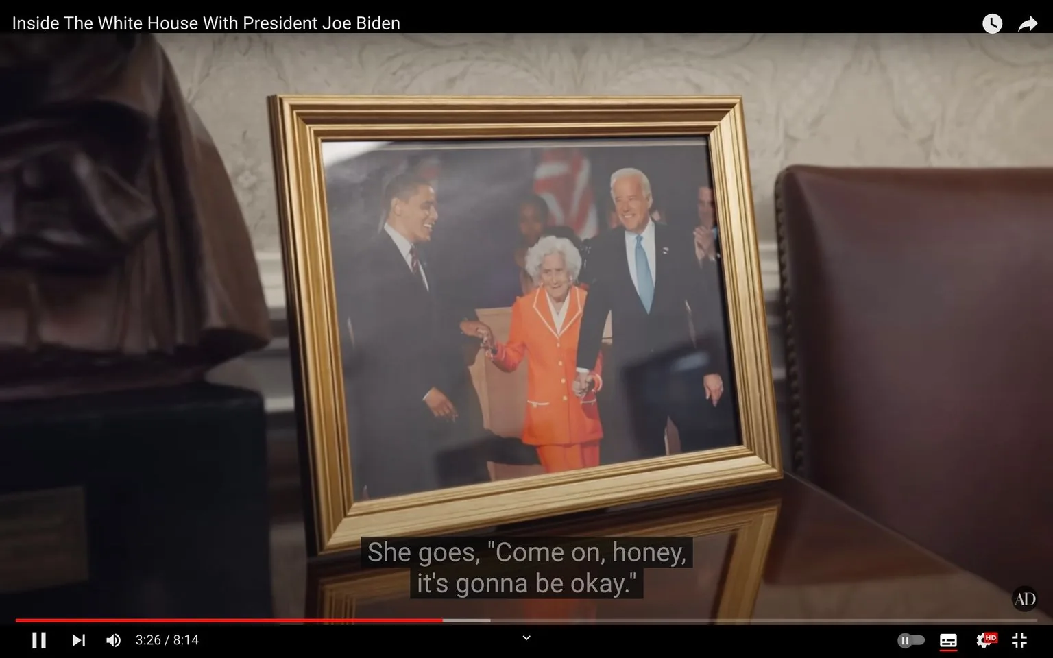 The image is a still frame from a YouTube video titled "Inside The White House With President Joe Biden." It shows a close-up of a framed photograph placed on a desk or table in an indoor setting, likely within the White House. The photograph features three individuals, including President Joe Biden and a woman in an orange outfit. The hint mentions Obama, suggesting his presence in the image. The frame is ornate and gold-colored, resting on a polished wooden surface. The background of the scene includes a decorative wall and part of a leather chair. The lighting is warm, highlighting the framed photo prominently. The image captures a moment of camaraderie and warmth, as suggested by the visible smiles and the descriptive caption, "She goes, 'Come on, honey, it's gonna be okay.'" This provides a sense of reassurance and personal connection within the larger context of the video.