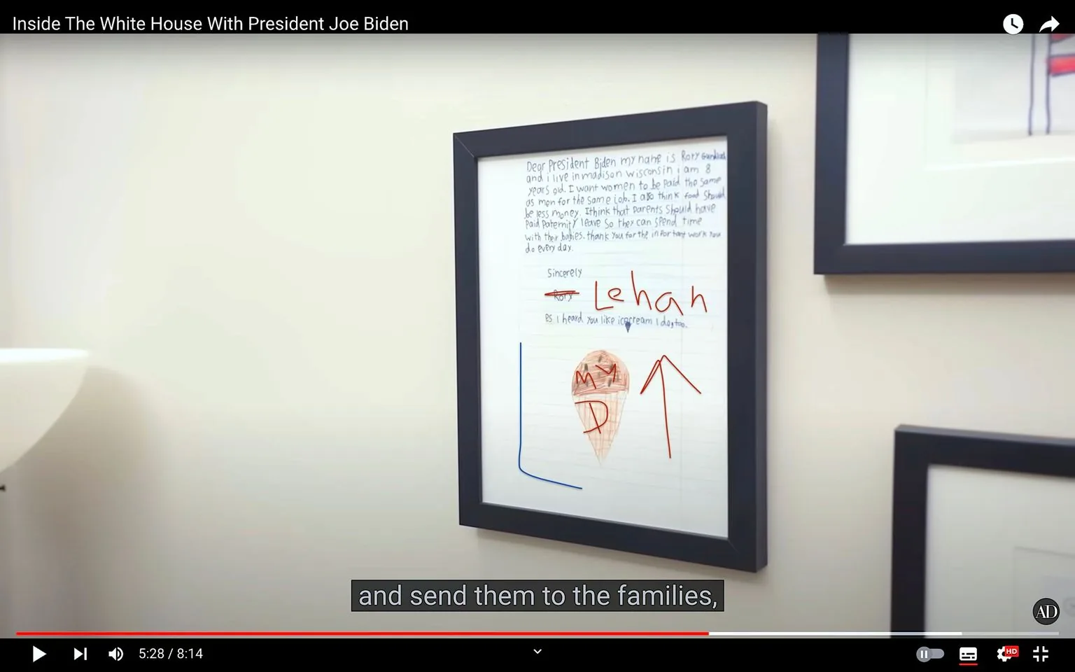 The image is a video frame from a YouTube video titled "Inside The White House With President Joe Biden." It features a close-up of a framed letter hanging on a light-colored wall. The letter is handwritten and appears to be from a child, addressed to President Joe Biden. The text in the letter expresses a desire for gender equality in pay and mentions parental leave. The child's name, "Lehan," is written in large, colorful letters at the bottom of the letter. An arrow and a small drawing, possibly depicting an ice cream cone, are also visible in the letter. The frame and the wall are simple and unadorned, creating a focus on the content of the letter. The setting suggests a personal or office space, possibly within the White House. The video is accompanied by captions, with one line visible: "and send them to the families," suggesting a discussion of family-related topics.