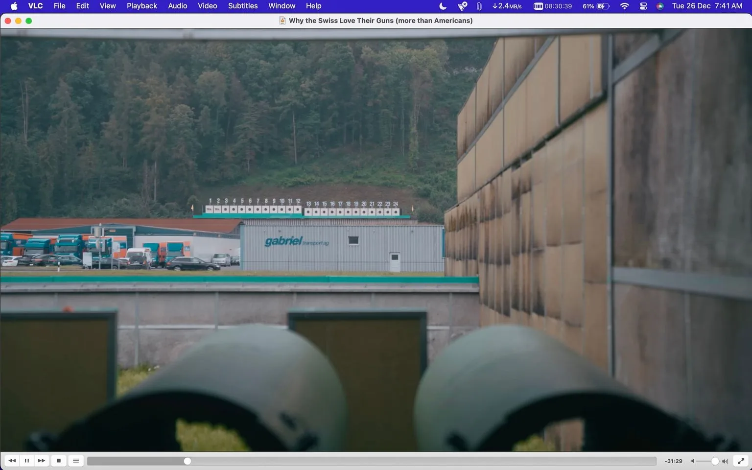 ```
The image is a screenshot from VLC media player, displaying a paused video titled "Why the Swiss Love Their Guns (more than Americans)". The scene is set outdoors, likely in a rural area, with a focus on a building labeled "gabriel transport ag". The building is situated near a row of trucks and vehicles, suggesting a logistics or transport hub. In the background, there's a forested hillside, adding to the rural atmosphere.
The foreground features part of a structure, possibly related to the transportation facility, with two rounded objects partially visible, which might be barrels or similar equipment. The lighting is natural, indicating daytime, and the overall color palette includes greens and browns, emphasizing the natural surroundings.
The screenshot's visible interface shows it's paused at 31:29, with a blue-themed VLC player control bar at the top. There's no specific channel or profile information visible within the image. Additionally, no captions or subtitles are present, and the forested hill and transport facility create a serene yet industrious setting.