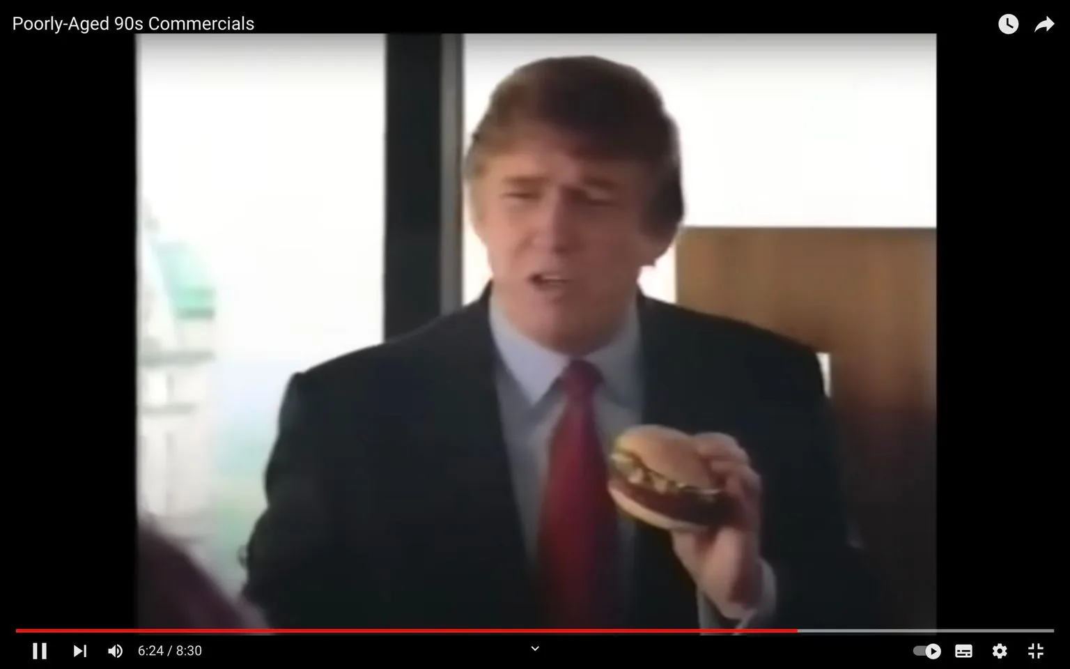 In this video frame from a YouTube video titled "Poorly-Aged 90s Commercials," Donald Trump is featured prominently. He is indoors, seated in what appears to be a corporate or upscale environment. Donald Trump is holding a hamburger in his hand, suggesting this is an advertisement likely related to fast food. He is wearing a dark suit with a white shirt and a red tie, characteristic of a formal business setting. His expression is animated, possibly mid-speech as he might be discussing the product he is holding. There is a building visible through the window in the background, hinting at an urban setting. The lighting is bright, indicating it's likely daytime. The frame captures a moment that reflects 1990s advertising styles, focusing on celebrity endorsements. The video, hosted on YouTube, is part of a compilation showcasing commercials from that era.