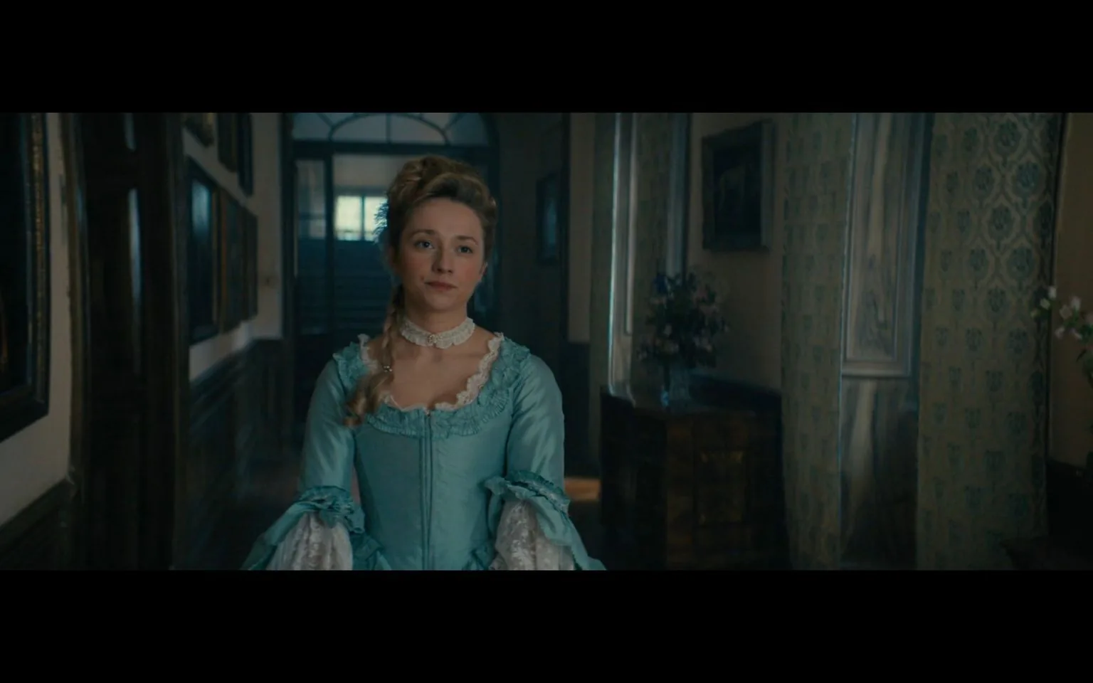 The image is a frame from the series "La Révolution." It features a woman dressed in an elaborate 18th-century style gown, predominantly light blue with white lace details. Her hairstyle is elaborate, with hair pulled back and styled high, typical of the period. She has a composed and serene expression, standing in a dimly lit hallway. The background includes dark wood paneling and framed paintings lining the walls, suggesting a setting in a grand, historical mansion. The lighting is soft, casting a warm and slightly muted tone over the scene, enhancing the historical ambiance. There is also a vase with flowers visible on a side table, adding a decorative touch to the hallway. The overall composition and costume design are consistent with the show's period setting, focusing on themes of historical drama.