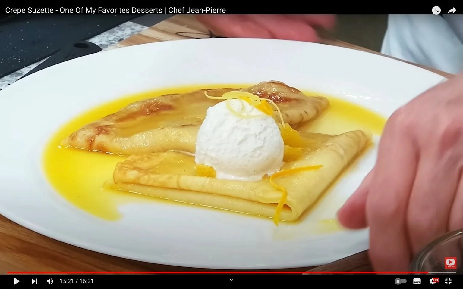 The video frame shows a close-up of a beautifully plated Crepe Suzette prepared by Chef Jean-Pierre. The dish features thin crepes elegantly folded and arranged on a white plate. A generous scoop of ice cream sits atop the crepes, while a vibrant yellow-orange sauce, likely a citrus reduction, surrounds them. Thin strips of orange zest are artfully placed on top, adding a touch of color and texture. The hands of Chef Jean-Pierre are visible, carefully positioning the plate, indicating meticulous attention to detail. The setting is a kitchen, with a wooden cutting board partially visible in the background, suggesting a cooking demonstration atmosphere. The lighting is bright, highlighting the rich colors and textures of the dessert. The video is titled "Crepe Suzette - One Of My Favorites Desserts | Chef Jean-Pierre" and is found on Chef Jean-Pierre's YouTube channel.