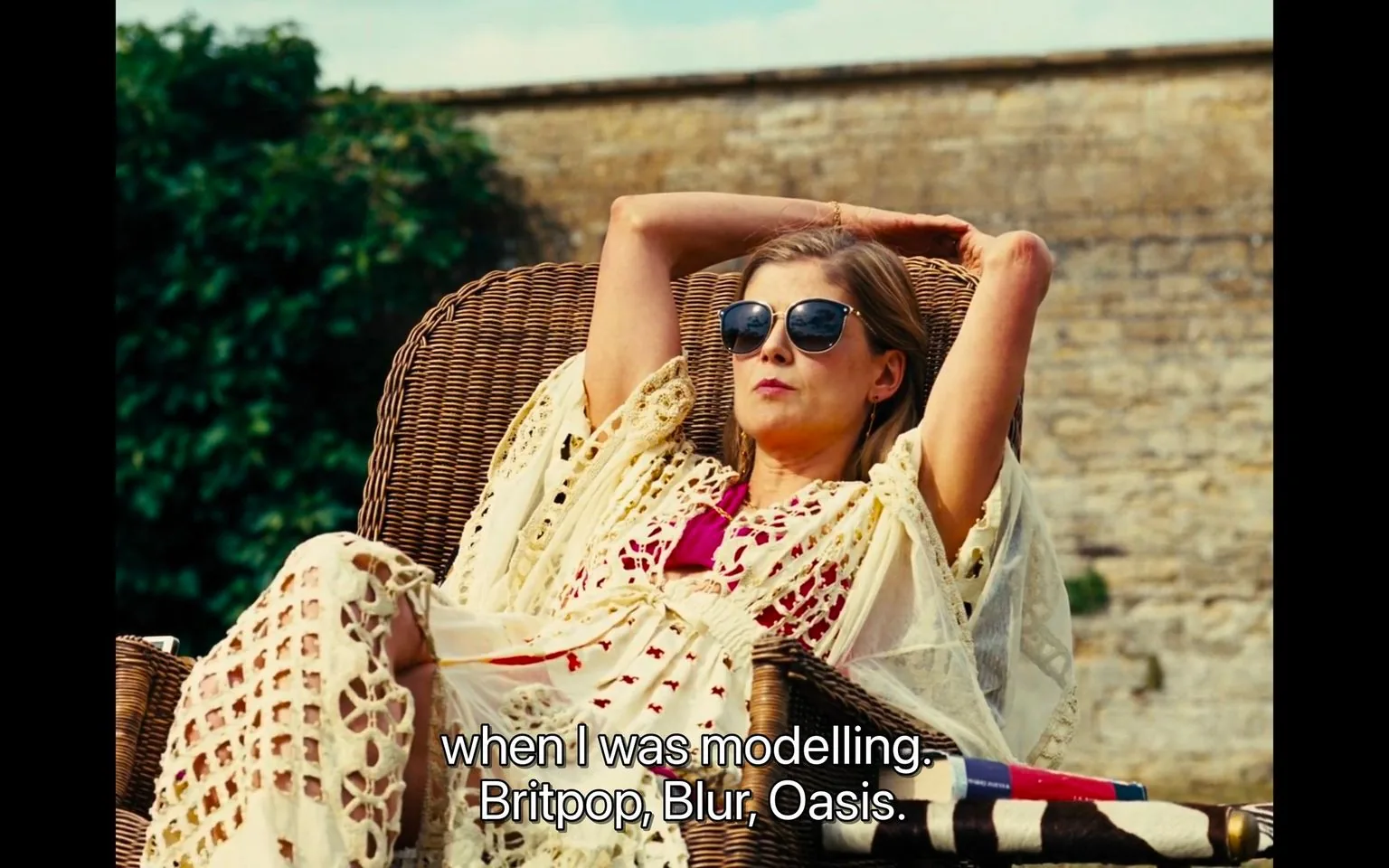The scene shows a woman lounging outdoors, seated comfortably on a wicker chair. She is wearing sunglasses and a cream-colored crochet cover-up over a bright pink outfit, suggesting a summery and relaxed atmosphere. Her arms are stretched behind her head as she leans back, exuding a laid-back demeanor. The background consists of a stone wall and some lush greenery, indicating a garden or backyard setting. The lighting is bright and warm, typical of a sunny afternoon.
The subtitles reveal she is reminiscing about her modeling days, referencing cultural elements like Britpop, and bands like Blur and Oasis, which adds a nostalgic tone to the scene. This information hints at a discussion or reflection on past experiences in the fashion industry during the era when those bands were prominent.