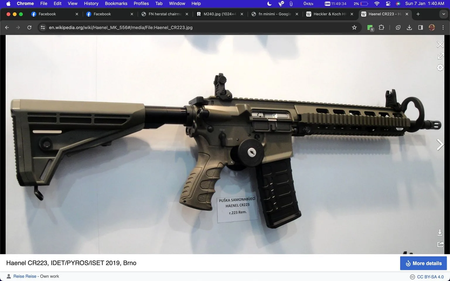 The image depicts a Haenel CR223 rifle displayed at IDET/PYROS/ISET 2019 in Brno. The rifle is mounted against a plain background, showcasing its design and features. It has a tactical appearance with a matte black finish and a collapsible stock. The rifle has a rail system on the upper receiver, allowing for the attachment of various accessories. The magazine is inserted, and the safety selector is visible near the grip. A tag hanging from the trigger guard provides information about the model and caliber: "PUŠKA SAMONABÍJECÍ HAENEL CR223 r .223 Rem." The image has a clean and professional look, typical of trade show displays, highlighting the technical and aesthetic aspects of the firearm.