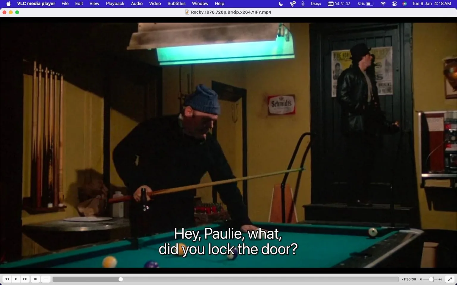 The image is a screenshot from the movie "Rocky" (1976), showing a scene inside a dimly lit bar or pool hall. The setting is characterized by a pool table in the foreground with pool balls scattered on it. Two men are present: one, wearing a beanie and dark clothing, is holding a pool cue and leaning over the table, possibly lining up a shot. He appears focused on the game. The other man is standing near a door in the background; he's dressed in a leather jacket and appears to be engaging in a conversation, as suggested by the subtitle: “Hey, Paulie, what, did you lock the door?”
The lighting is subdued, with a green overhead lamp providing most of the illumination directly above the pool table. There are pool cues stored neatly in the background. A small sign reading "Schmidt's" can be seen on the wall, indicating a brand or advertisement. The atmosphere is intimate and somewhat tense, suggesting a private or important conversation amidst a casual game of pool. The media player interface visible at the top of the image shows it's being played on VLC, with controls and a timeline indicating the video is at 1 hour and 36 minutes into the film.