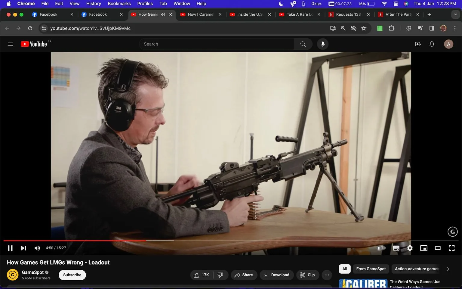 The image is a screenshot from a YouTube video titled "How Games Get LMGs Wrong - Loadout" on the GameSpot channel. The scene takes place indoors, likely in a studio or demonstration setting. A man is seated at a wooden table, wearing protective earmuffs and safety glasses. He appears engaged in examining or discussing a light machine gun (LMG) placed on the table in front of him. The firearm is mounted on a bipod, and there is a coil of ammunition visible beneath it. The background features a simple, neutral setup with some metal framework visible. The lighting is bright and even, suggesting a controlled environment typical of a recording studio. The video seems to be focusing on the accuracy of LMGs in video games versus real life.