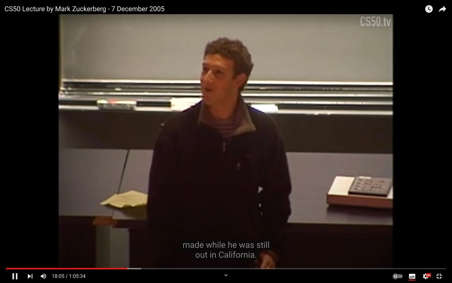 In this video frame, Mark Zuckerberg is giving a lecture as part of the CS50 course on December 7, 2005. He is positioned at the front of a classroom, likely at a university setting, as suggested by the lecture environment. The room features a chalkboard behind him and a desk with papers and possibly a book or electronic device resting on it. Zuckerberg is dressed casually in a dark jacket over a striped shirt, and his posture is relaxed as he looks slightly to his left, suggesting engagement with an audience or looking towards a screen or board. The lighting is bright enough to illuminate him clearly against the darker background of the classroom. Subtitles from the video indicate he is discussing something related to "California," hinting at the context of his speech. The lecture likely covers topics related to technology or business, given Zuckerberg's background and the educational setting.
- Title: "CS50 Lecture by Mark Zuckerberg - 7 December 2005"
- Channel / profile: "CS50.tv"
- Site / app: "YouTube"
- Captions / subtitles: "made while he was still out in California."
