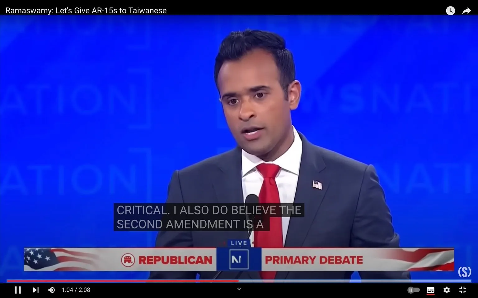 Vivek Ramaswarmy is featured in this video frame taken from a Republican Primary Debate. He is positioned in the foreground, speaking into a microphone. Ramaswarmy is wearing a dark suit with a white shirt and a red tie, with a small American flag pin on his lapel, indicating his participation in a formal political setting. The background is a solid blue with indistinct patterns, suggesting a large event or stage backdrop typical of televised debates.
The title of the video, "Ramaswamy: Let's Give AR-15s to Taiwanese," appears at the top, implying a controversial or significant statement made during the debate. The on-screen text includes a subtitle that reads, "CRITICAL. I ALSO DO BELIEVE THE SECOND AMENDMENT IS A," suggesting a discussion related to gun rights or foreign policy. Below Ramaswarmy, a banner with the text "REPUBLICAN PRIMARY DEBATE" is prominently displayed, adorned with an American flag motif. The overall mood of the scene is serious and focused, reflecting the formal and high-stakes nature of political debates.