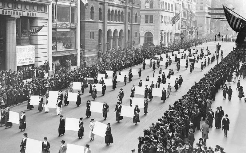 This black-and-white photograph captures a historical parade scene on a wide city street lined with buildings and large crowds on both sides. In the center, a procession of individuals, predominantly women, march in orderly rows holding blank placards. The participants are dressed in early 20th-century attire, with long coats, hats, and skirts, conveying a formal and organized appearance. The street is flanked by tall buildings, including one with a sign reading "NATIONAL BANK" and another promoting "LIBERTY BONDS" with the phrase "BE A PATRIOT."
The street is bustling with onlookers, tightly packed along the sidewalks, watching the parade with interest. The atmosphere is one of solemnity and purpose, suggesting a significant social or political event. Flags and banners adorn the buildings, contributing to the sense of occasion. The lighting is bright and even, typical of an overcast day, which adds to the timeless quality of the photograph. This image is a vivid snapshot of a moment in history, capturing the spirit and determination of the participants.