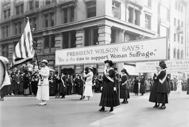 The image depicts a historical scene from an early 20th-century suffrage parade advocating for women's right to vote. Prominently featured is a large banner that reads, "President Wilson Says: This is the time to support Woman Suffrage," highlighting the support of President Wilson for the cause. The parade takes place on a city street lined with tall, classical-style buildings, suggesting an urban setting.
In the foreground, several women are marching in formation. They are dressed in period attire typical of the early 1900s, with long skirts and hats. Some women wear sashes, possibly indicating their roles as leaders or organizers within the suffrage movement. The mood is solemn and determined, reflecting the serious nature of their cause.
The crowd gathered along the street watches the march, emphasizing the public interest and support that the movement garnered. The black-and-white coloration of the image gives it an authentic historical feel, capturing a significant moment in the fight for women's rights. The lighting is natural, suggesting a daytime event, and the overall composition conveys the sense of unity and purpose among the participants.
- Title: "President Wilson Says: This is the time to support Woman Suffrage."