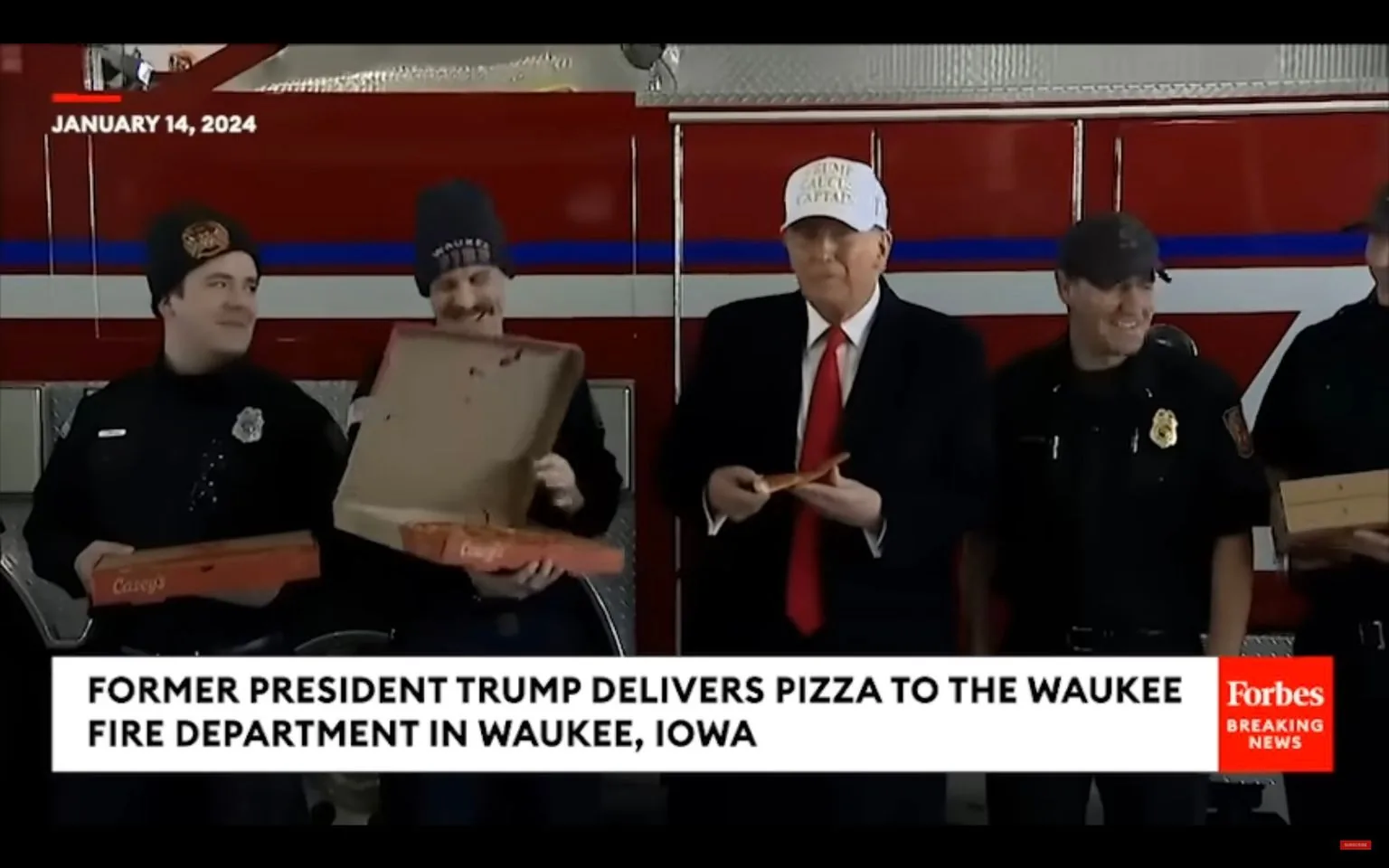 The image shows Donald Trump, identified by the hint as "Trump," delivering pizza to members of the Waukee Fire Department in Waukee, Iowa. The setting is indoors, likely inside the fire station, as indicated by the red fire truck in the background. Trump is wearing a white hat that says "TRUMP" and a suit with a red tie, holding a slice of pizza. Next to him are several fire department personnel dressed in black uniforms, some wearing caps. One of the firefighters is holding an open pizza box, smiling, while others seem to be enjoying the moment, creating a cheerful atmosphere. The lighting is even and bright, typical of an indoor event setting. The image captures a moment of interaction and appreciation, with Trump participating in a community service gesture. The Forbes Breaking News logo is visible, indicating the source of the coverage.
- Title: "FORMER PRESIDENT TRUMP DELIVERS PIZZA TO THE WAUKEE FIRE DEPARTMENT IN WAUKEE, IOWA"
- Publisher / studio: "Forbes Breaking News"
- Key OCR: "JANUARY 14, 2024"