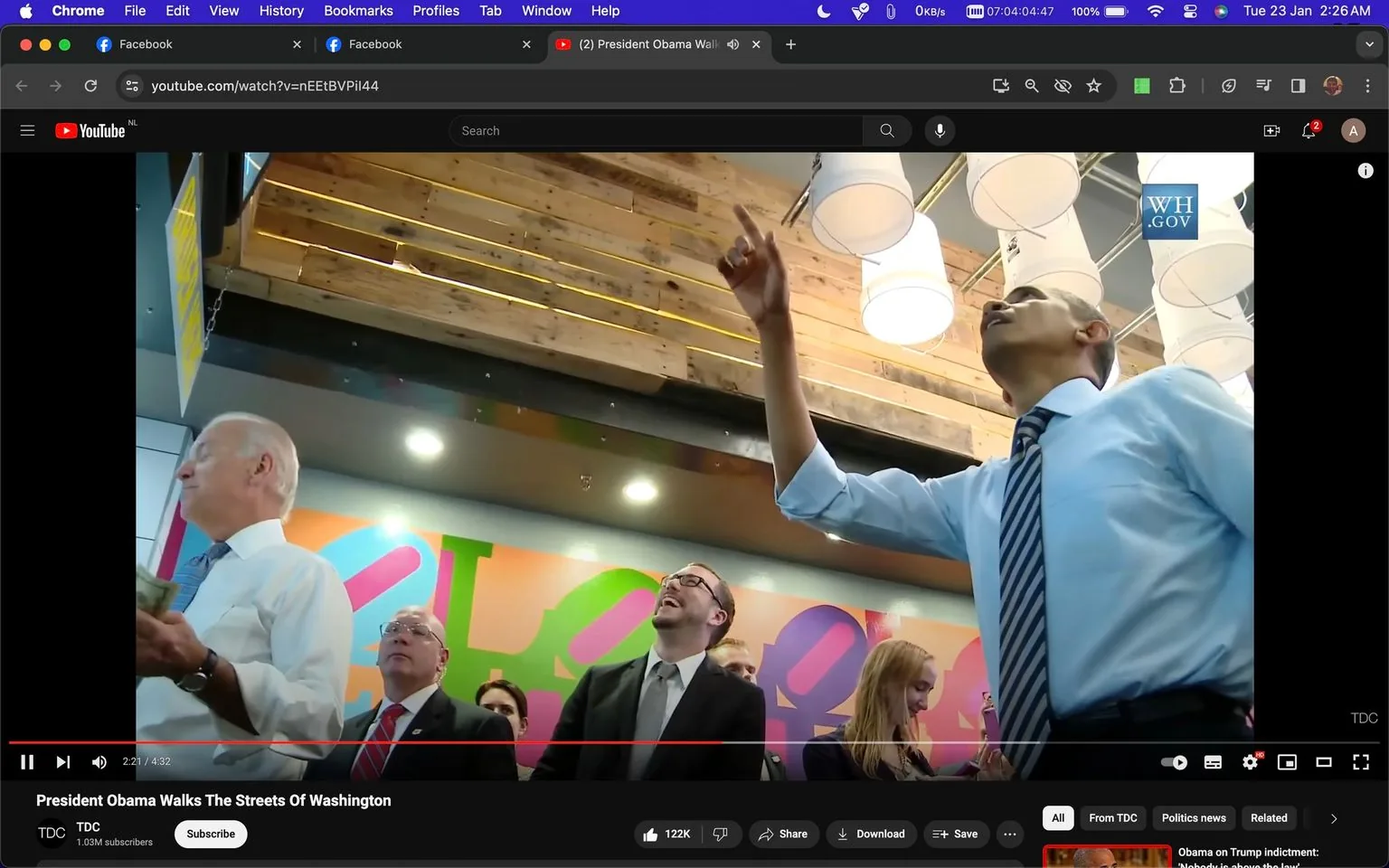 President Barack Obama and Joe Biden are featured in this YouTube screenshot titled "President Obama Walks The Streets Of Washington," captured from the channel TDC. They are inside a colorful and modern setting, possibly a cafe or public venue. Barack Obama is in the foreground, looking upwards and gesturing with his right hand, implying he could be explaining or pointing something out. He is dressed in a light blue shirt with rolled-up sleeves and a striped tie, giving a casual yet professional appearance. Joe Biden is visible to the left, holding what looks like money in his hand, suggesting a transaction or purchase. He appears engaged, looking in the same direction as Obama. The background is lively with various people, some smiling and appearing attentive. The lighting is bright with ceiling lamps visible, and a colorful mural adorns the wall, adding vibrancy to the scene. The screenshot is part of a video uploaded to YouTube, as indicated by the URL and platform branding present in the image.