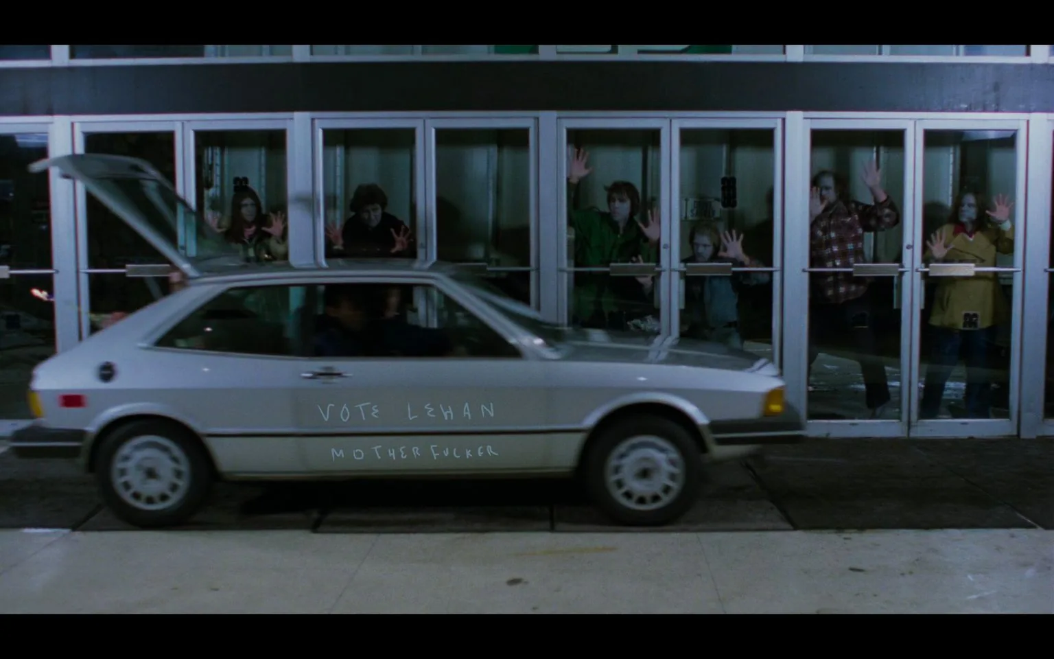 The image depicts a scene from the film "Dawn of the Dead." In the foreground, a silver car is parked with its hatchback open. The side of the car has graffiti that reads "VOTE LEHAN MOTHERFUCKER." Visible through the car windows is a person inside, though their identity is unclear. In the background, several individuals, who appear to be zombies, press their hands against large glass doors, creating a barrier between themselves and the outside. They exhibit the typical disheveled appearance and vacant expressions associated with zombies. The scene is set at night, with artificial lighting illuminating the area, casting reflections on the glass. The setting appears to be the entrance of a mall or large building, contributing to the atmosphere of a classic zombie apocalypse.