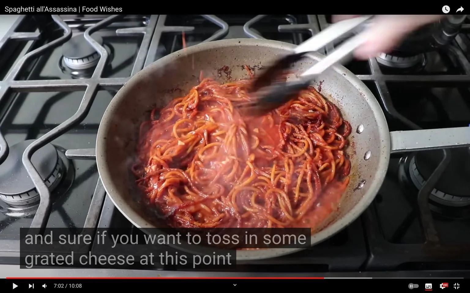 The video frame shows a cooking scene from the YouTube video titled "Spaghetti all'Assassina | Food Wishes" on the Food Wishes channel. The setting is a kitchen, specifically focused on a stovetop where a pan of spaghetti is being cooked. The pasta is visibly coated in a rich, red sauce, typical of tomato-based dishes. It seems to be in the process of being tossed with tongs, suggesting active cooking. The stovetop is modern with several burners visible, and the focus is on the pan which holds the richly colored spaghetti. The lighting is bright, highlighting the steam rising from the dish, adding an element of warmth and freshness. The color palette is dominated by the deep red of the sauce, contrasted with the metallic tones of the pan and the black of the stove grates. The accompanying subtitles suggest a casual, conversational tone as the narrator discusses the option of adding grated cheese. The scene captures the essence of home cooking with an emphasis on Italian-inspired cuisine.
- Title: "Spaghetti all'Assassina | Food Wishes"
- Channel / profile: "Food Wishes"
- Site / app: "YouTube"
- Captions / subtitles: "and sure if you want to toss in some grated cheese at this point"