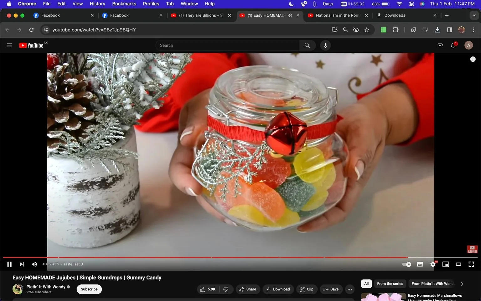 The screenshot is from a YouTube video titled "Easy HOMEMADE Jujubes | Simple Gumdrops | Gummy Candy" on the channel "Platin' It With Wendy." The image shows a close-up of a pair of hands holding a decorative glass jar filled with colorful homemade jujubes or gummy candies. The jar is adorned with a red ribbon and a shiny red bell, giving it a festive appearance. Next to the jar, there's a decorative vase containing snowy pinecones and frosted evergreen branches, enhancing the holiday theme. The hands are carefully presenting the jar, suggesting a warm, inviting atmosphere. The video timeline shows a duration of 4:15/4:59, indicating the video is nearing the end. The overall setting is cozy and festive, suitable for a holiday-themed candy-making tutorial. The browser tabs at the top display open tabs for Facebook, and "They are Billions," alongside the YouTube video.