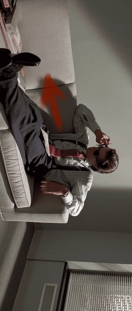 The image shows a man reclining on a sofa in an office setting. He is dressed in business attire, featuring a striped shirt, red suspenders, and dark trousers. The man is casually lounging with one leg stretched out on the sofa and the other bent, while he appears to be talking on a phone, adding a relaxed yet engaged demeanor to the scene. His sunglasses add a touch of nonchalance.
The room is characterized by a simple design with neutral-colored walls and a carpeted floor. In the background, there is a window with blinds partially drawn, allowing some light to filter into the room. A visible air conditioning unit is situated beneath the window. The overall lighting is subdued, creating a calm and understated atmosphere.
An arrow is drawn on the sofa, pointing towards the man, possibly indicating a humorous or significant aspect of the scene. The composition suggests a moment of casual conversation or reflection. The overall mood combines elements of professionalism with a hint of irreverence, fitting the context of the named work "The Unknown Known" and the playful title "Peekaboo!!!!!!".