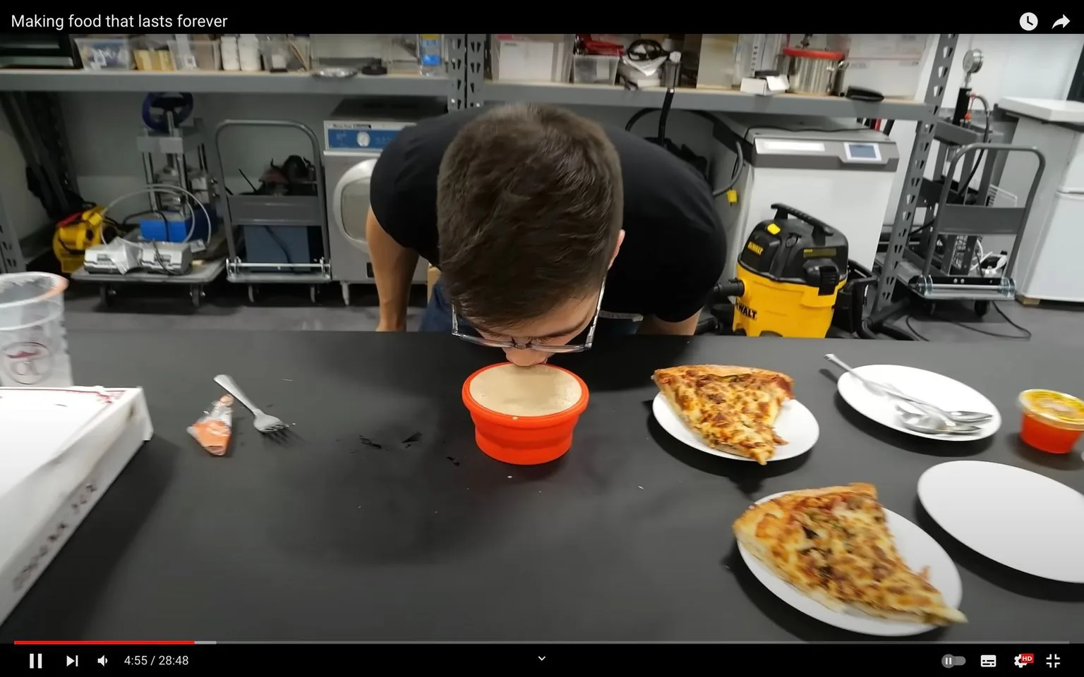 The image is a screenshot from a video titled "Making food that lasts forever." It shows a person leaning over a table in what appears to be a laboratory or workshop setting. The individual is drinking directly from a red bowl placed on the table. On the table, there are two plates each with a slice of pizza, and some utensils nearby. In the background, shelves with various equipment and a yellow vacuum cleaner are visible. The setting suggests an experimental or casual scientific environment focused on food preservation or longevity. The overall mood is casual, likely involving some form of food experiment as indicated by the video title. The lighting is bright and evenly distributed, highlighting the person's action in the center of the frame.