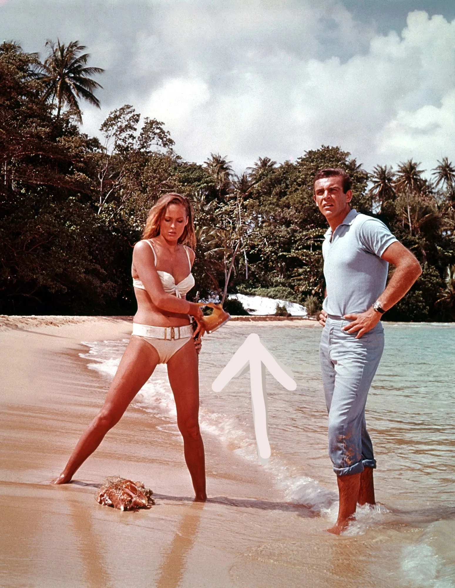 The image captures a classic scene from the film "Dr. No," showcasing Sean Connery on a sandy beach. Sean Connery, wearing a light blue polo shirt and rolled-up light blue pants, stands on the shoreline with his hands on his hips. Nearby, a woman in a white bikini holds an object in her hand, with a large conch shell at her feet. The lush green foliage and palm trees in the background frame the setting, highlighting the tropical environment. The sky is partly cloudy, casting a soft, natural light over the scene, suggesting a warm, sunny day. This iconic moment emphasizes the adventurous and exotic themes typical of the James Bond series, with Sean Connery's confident posture reflecting his character's suave and fearless demeanor.