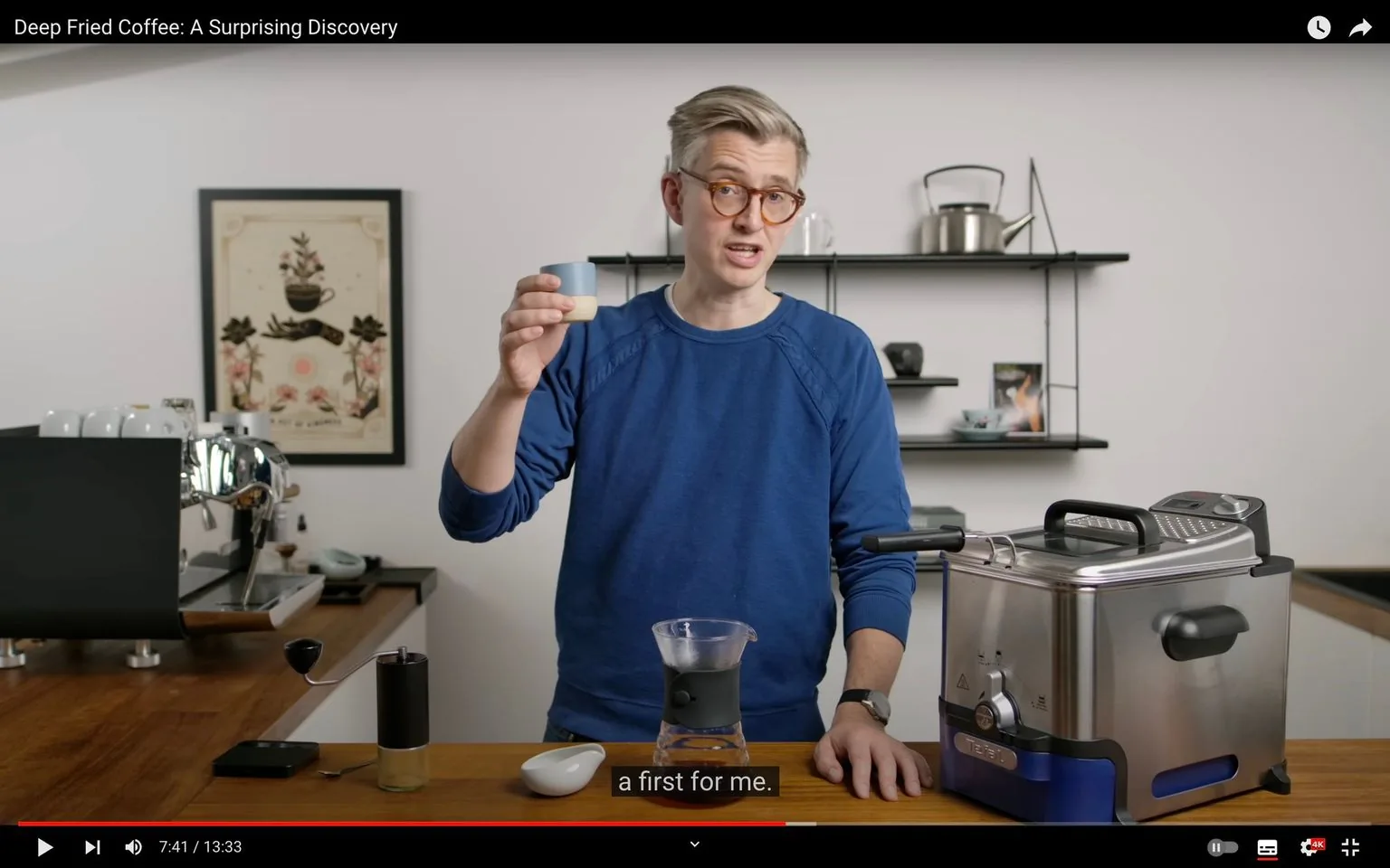 James Hoffmann is shown in a video frame from his YouTube channel. He stands in a modern kitchen setting, engaging in a demonstration. James is wearing a blue sweatshirt and glasses, holding a small cup in one hand as he explains something, indicated by the subtitle "a first for me." The setting is well-lit, with a clean and organized appearance. To his right is a deep fryer, suggesting that he might be talking about a coffee experiment involving deep frying. Behind him, shelves display kitchen items including a kettle and some cups, while a framed picture featuring a coffee theme decorates the wall. The countertop in front of him holds various coffee-making tools, including a coffee grinder. The scene conveys a sense of curiosity and exploration in coffee brewing methods.
- Title: "Deep Fried Coffee: A Surprising Discovery"
- Channel / profile: "James Hoffmann"
- Captions / subtitles: "a first for me."
- Site / app: "YouTube"