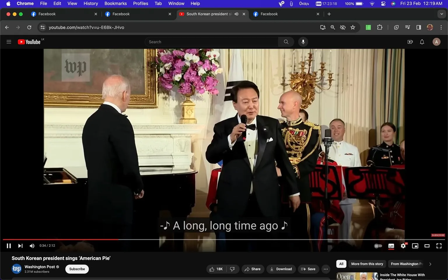 The image is a screenshot from a YouTube video titled "South Korean president sings ‘American Pie’," published by the Washington Post. The scene takes place indoors, likely at a formal event or gathering. In the foreground, a man with a microphone is dressed in a black tuxedo with a white shirt and bow tie, suggesting a formal occasion. Behind him stand several individuals in military uniforms, indicating a ceremonial or official atmosphere. A large floral arrangement and ornate decor are visible, adding to the formal setting.
In the left part of the image, another man, whose back is to the camera, seems to be listening to the singer. The setting is well-lit, with warm lighting reflecting off the white walls and elegant furnishings. A piano is partially visible, hinting at the musical nature of the event. The subtitle on screen reads, "A long, long time ago," a line from the song "American Pie," which the South Korean president is performing. The mood appears to be celebratory and engaging, as evidenced by the attention of the audience members.
- Title: "South Korean president sings ‘American Pie’"
- Channel / profile: "Washington Post"
- Site / app: "YouTube"
- Captions / subtitles: "A long, long time ago"
