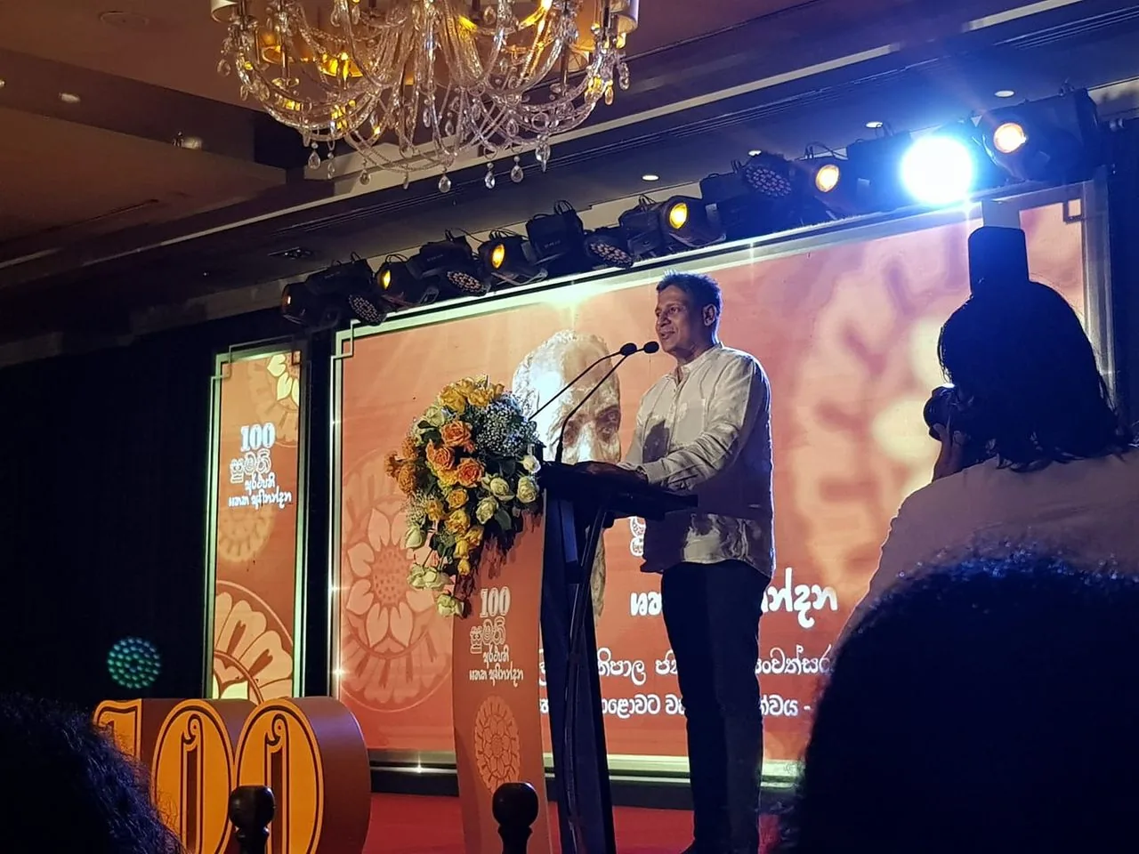 The image captures an indoor event featuring Dilith Jayaweera speaking at a podium. He is dressed in a white shirt and is positioned under a bright stage light, enhancing the focus on him. The backdrop is a warm, orange-toned screen with floral patterns and the number "100" prominently displayed, suggesting a centennial celebration or significant milestone event. A large bouquet of flowers decorates the podium, adding a touch of elegance. The setting includes a chandelier hanging from the ceiling, contributing to a formal atmosphere. An audience is present, and a person is seen taking photographs, indicating media coverage. The overall mood is celebratory and formal, with well-coordinated lighting and decor. The presence of professional photography equipment highlights the importance of the occasion.