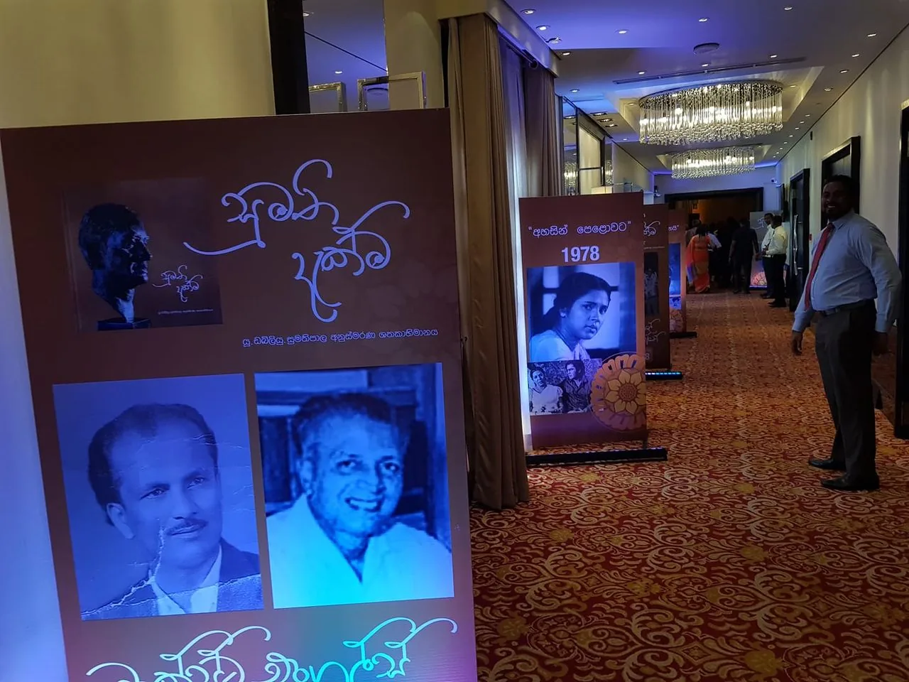 The image is set in a well-lit hallway adorned with red and gold patterned carpet, suggesting a formal event or exhibition. On the left side, there's a large display featuring photographs and text. The hint identifies "U W Sumathipala" as the person in the left picture. The display has a warm brown background with elegant script and includes two black-and-white portraits under the main text.
Further down the hallway, another panel displays a black-and-white photograph labeled "1978." The exhibition seems to commemorate or celebrate historical figures or events from Sri Lanka, as suggested by the text and style. Attendees in formal attire are visible in the background, engaging with the exhibits. The ambience is bright, with overhead lighting and a chandelier providing illumination. The overall mood is respectful and reminiscent, highlighting the importance of the individuals or events being showcased.
- Title: "U. W. Sumathipala Prathibha Abhimani Sanhinda"
- Named Works: "1978"