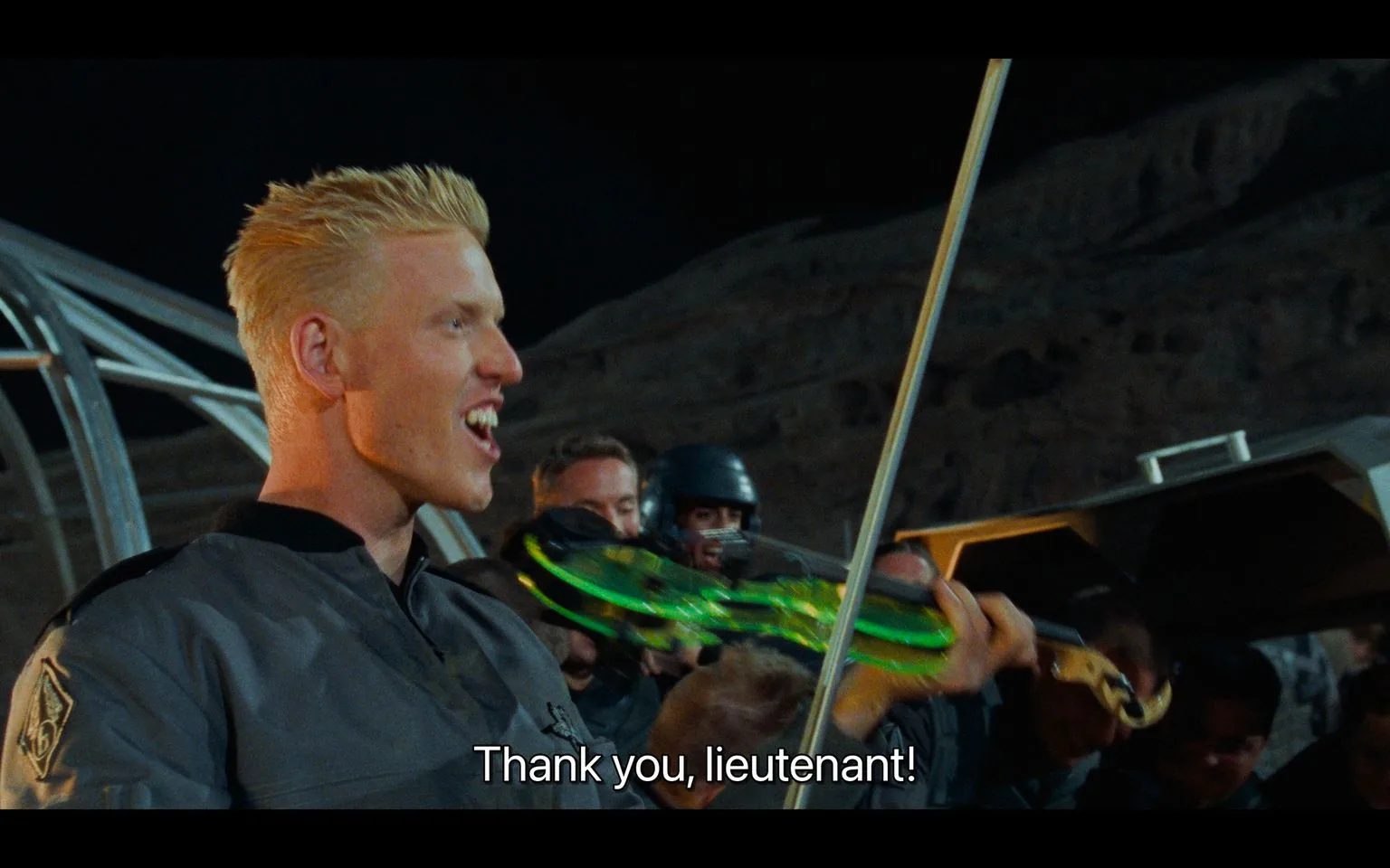 In this scene from "Starship Troopers," a character with short blonde hair is in focus, holding a violin-like object glowing with green light. He is wearing a military-style uniform, which indicates his association with a futuristic military setting. The environment suggests an outdoor, night-time celebration, with dim lighting that highlights the character's enthusiastic expression. The background shows several other people, some wearing helmets, appearing to join in the festivities.
The scene captures a moment of camaraderie and relief, possibly following a successful mission, as indicated by the character's grateful words, "Thank you, lieutenant!" The overall mood is upbeat and energetic, with the characters seemingly enjoying a rare moment of joy amidst the challenges they face. The visual style is typical of late 1990s sci-fi films, featuring futuristic elements and a mix of practical and special effects. The film's themes include military life, teamwork, and the struggles against alien adversaries.