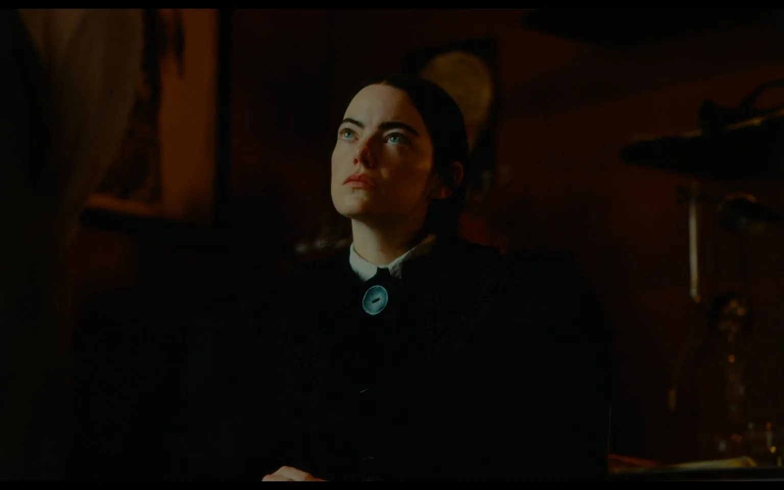 Emma Stone is depicted in this scene from the film "Poor Things." She sits indoors, illuminated by dramatic, subdued lighting that casts shadows across her face, enhancing the intensity of her gaze. Her expression is thoughtful and slightly melancholic, as she looks slightly upward, possibly at another character or an object out of the frame. She wears period attire, consisting of dark clothing with a prominent, circular blue brooch at her collar, which contrasts against her otherwise somber outfit.
The background is dimly lit, featuring indistinct objects that suggest a setting rich in detail, possibly a study or a parlor typical of a bygone era. The composition is intimate, focusing on Emma Stone's character, drawing attention to her nuanced expression and the subtle textures of her clothing. The overall mood is contemplative and introspective, suggestive of a moment of introspection or realization within the narrative.