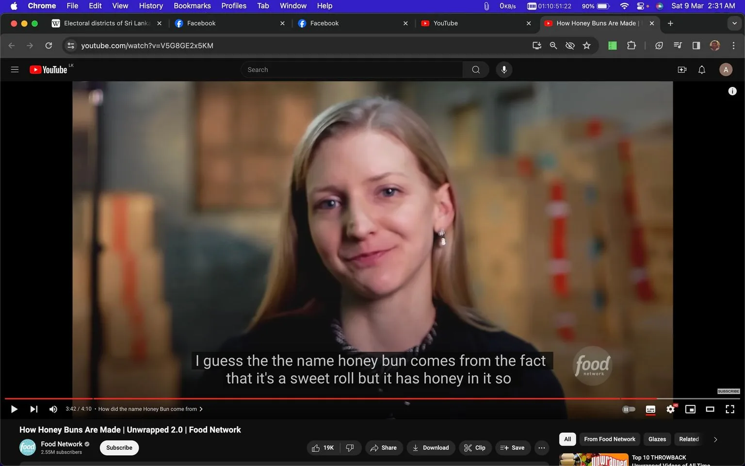 The screenshot is from a YouTube video titled "How Honey Buns Are Made | Unwrapped 2.0 | Food Network," uploaded by the Food Network channel. The video appears to be part of the series "Unwrapped 2.0," which explores the making of various foods. In the image, a woman with long blonde hair and earrings is speaking in an interview setting. She is smiling slightly and looking directly at the camera, suggesting a conversational tone. The background is softly blurred, with stacks of boxes visible, indicating an industrial or bakery environment. The lighting is warm, casting a natural and welcoming ambiance. The Food Network logo is visible in the lower right corner. Subtitles at the bottom of the screen read, "I guess the the name honey bun comes from the fact that it's a sweet roll but it has honey in it so," providing context about honey buns. The interface shows typical YouTube elements like play controls and a video timeline at the bottom.