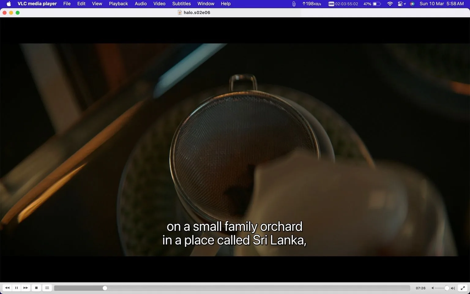 The screenshot depicts a scene from the series "Halo" being played on VLC media player. The interface is visible, showcasing typical playback controls and a title bar indicating the file name: "halo.s02e06.2160p.web.h265-halohowareyou.mkv." The image captures a close-up view of a sieve being used to pour what appears to be a liquid, possibly tea or coffee, with a brownish tint. The lighting is warm and highlights the texture of the sieve.
Subtitles at the bottom of the image read: "on a small family orchard in a place called Sri Lanka," suggesting a narrative related to the origin of the beverage or an ingredient. The background and surroundings are softly blurred, focusing attention on the pouring action. This setting seems intimate, likely a kitchen or similar environment where such preparation takes place.