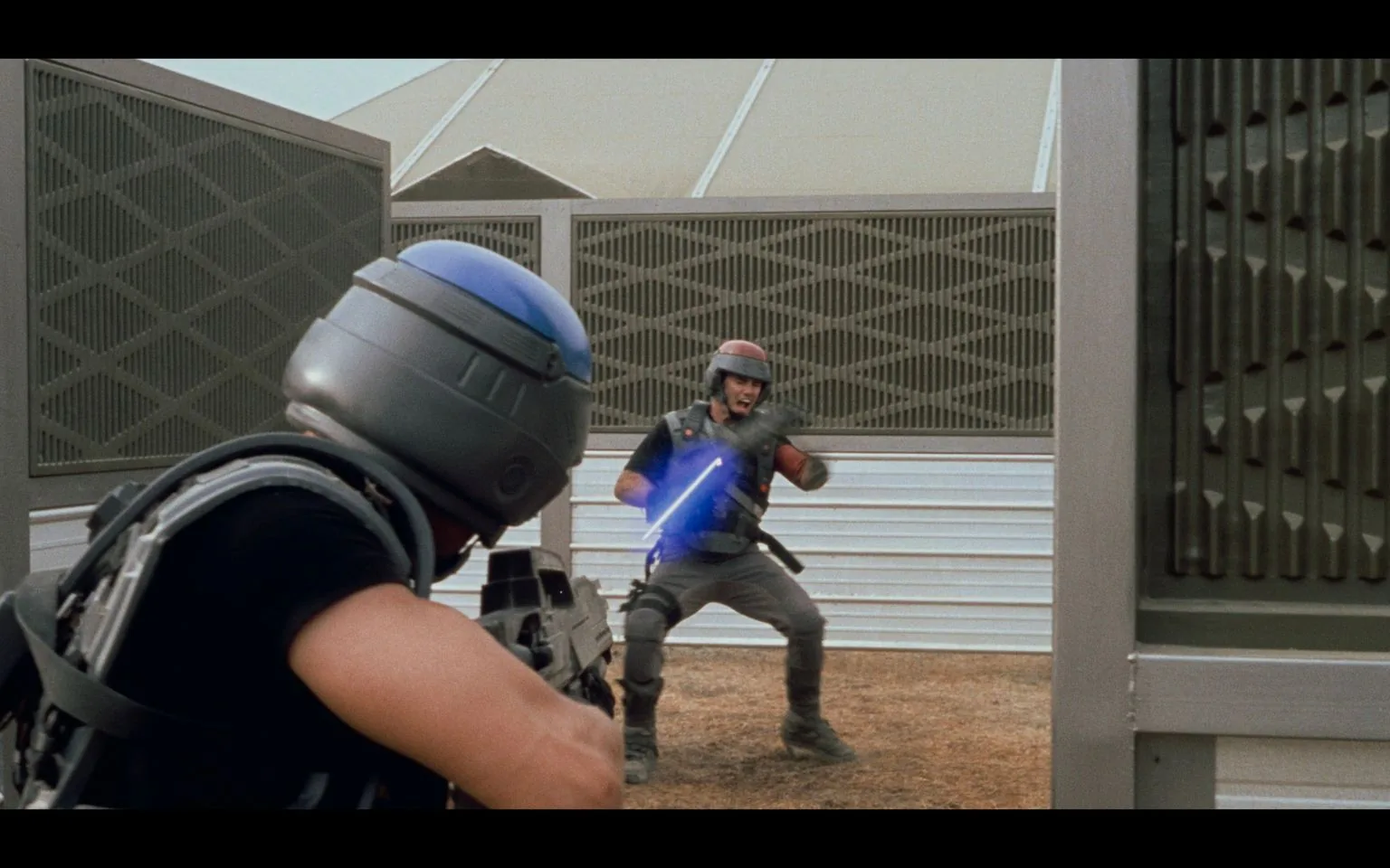 In this scene from "Starship Troopers," two characters are engaged in a tense combat situation. The setting appears to be a training or battle zone with futuristic architecture, including large metallic structures and barriers. In the foreground, one character is seen from behind, wearing a helmet with a blue visor and tactical gear. This character is holding a weapon aimed at the character in the background.
The character in the background is wearing a similar helmet with a red stripe and is reacting as if being shot, evidenced by a blue laser-like effect hitting them. This individual is also dressed in military attire, emphasizing the science fiction military theme of the film. The posture and expression of the background character suggest surprise or pain, adding to the dramatic tension of the scene. The colors are muted, with a focus on metallic grays and blues, consistent with the futuristic and militaristic style of "Starship Troopers." The lighting is natural, indicating an outdoor setting during daytime.