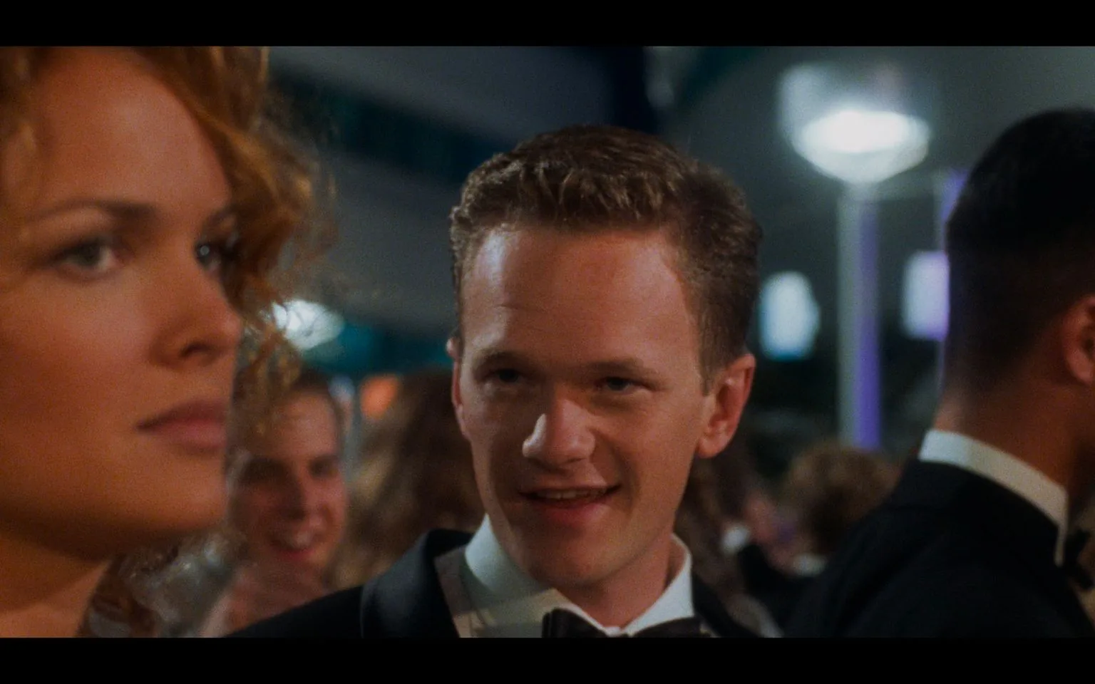 Neil Patrick Harris is depicted in this screenshot from the movie "Starship Troopers" (1997). The scene takes place at an indoor, formal event, likely a dance or ceremonial gathering. Neil Patrick Harris is wearing a classic black tuxedo with a bow tie, fitting the formal theme of the event. His expression is engaged and slightly amused, suggesting an interaction with someone nearby. The lighting is soft and ambient, characteristic of an evening event, with blurred figures and lights in the background that add to the atmosphere of a lively social gathering. The woman in the foreground, slightly out of focus, has curly hair and is also dressed formally, contributing to the festive setting. The mood of the scene is social and elegant, capturing a moment of interaction amidst a crowd.