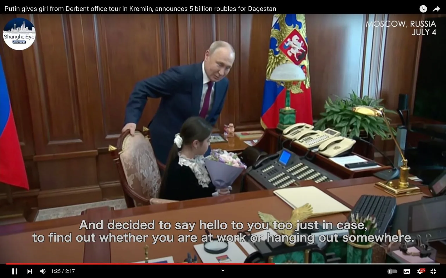 1) Rich description:
The scene is set in a formal office, likely part of the Kremlin, where a significant meeting is taking place. The center focus is a man, identified by the hint as Putin, who is interacting with a young girl seated at a large, polished desk. The girl is holding a bouquet of flowers, suggesting a ceremonial or welcoming gesture. Putin, dressed in a formal suit and tie, appears to be engaging warmly, leaning slightly toward the girl as if in conversation. The room is furnished with ornate, classic décor, featuring wooden paneling and a large desk equipped with multiple phones and a lamp. A Russian flag is prominently displayed in the background, alongside an emblematic banner. The atmosphere is formal yet congenial, highlighted by the presence of a child in such an official setting. The lighting is bright, casting an even glow across the room, emphasizing the opulent details. The video frame includes subtitles at the bottom, giving context to their conversation.
2) On-screen text block:
- Title: "Putin gives girl from Derbent office tour in Kremlin, announces 5 billion roubles for Dagestan"
- Channel / profile: "ShanghaiEye"
- Site / app: "YouTube"
- Captions / subtitles: "And decided to say hello to you too just in case," "to find out whether you are at work or hanging out somewhere."