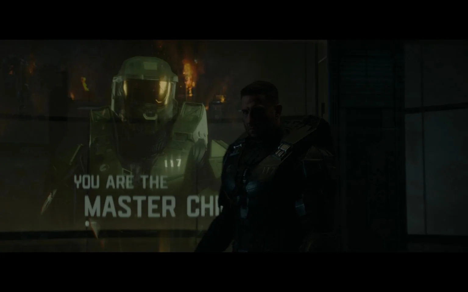 The image depicts a scene with a dark, intense atmosphere. In the foreground, a person is wearing futuristic armor, reminiscent of a soldier from a sci-fi setting. The armor is dark and has a tactical appearance, with the number "117" visible on the chest. This character is likely Master Chief, a well-known figure from the "Halo" series. In the background, a holographic image of a soldier in full armor, also identified as Master Chief, is prominently displayed. The hologram is bright, contrasting with the dimly lit room, and it includes the text "YOU ARE THE MASTER CHIEF." The setting suggests a military or futuristic environment, possibly involving a moment of reflection or briefing. The lighting is minimal, adding to the tension and seriousness of the scene.