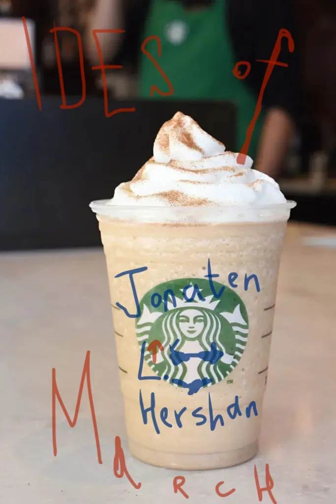 The image features a Starbucks coffee cup prominently placed in the foreground on a counter, likely within a cafe setting. The cup is filled with a blended beverage topped with whipped cream and sprinkled with cinnamon or cocoa powder. The iconic green Starbucks logo is clearly visible on the cup. Overlaying the image, text in red and blue reads "IDES of March" and "Jonaten Hershan." The text appears to have been digitally added, providing a humorous or thematic twist to the scene. In the background, an employee is partially visible, wearing the recognizable green apron associated with Starbucks staff, adding to the setting's authenticity. The lighting is bright, typical of a cozy cafe environment. The overall mood is lighthearted, possibly crafted for a humorous take on the "Ides of March" theme, juxtaposed with the everyday ritual of enjoying a coffee. The composition draws focus to the coffee cup and its customized text, creating an engaging visual narrative.
