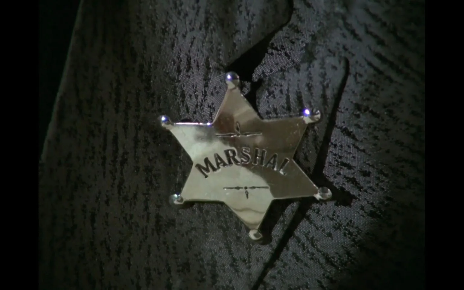 The image is a close-up shot of a shiny, silver star-shaped badge with the word "MARSHAL" engraved on it. This badge stands out against a textured, dark-colored fabric, likely part of a uniform or costume. The lighting highlights the metallic surface of the badge, creating reflections that draw attention to the engraved text. The scene is linked to "Living in Harmony," which suggests a context related to Western or law enforcement themes. The badge's design is simple yet iconic, reminiscent of traditional law enforcement insignia often seen in Western films or television shows. The overall mood is serious and authoritative, emphasizing the role of a marshal or law figure. The fabric in the background features a subtle pattern, adding depth and texture to the image.