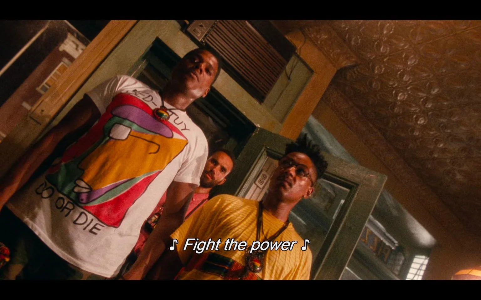 The image is a scene from the film "Do the Right Thing." It captures a dramatic moment indoors, with three men standing in close formation. The man in the foreground on the left wears a white T-shirt featuring a colorful abstract design, with the words "DO OR DIE" visible. The man in the center wears a yellow shirt, glasses, and a similarly styled necklace. Both men exhibit serious expressions, contributing to the intense atmosphere of the scene. In the background, a third man is partially visible, adding depth to the composition. The setting appears to be a room with ornate tin ceilings, suggesting a location with rich texture and history. The lighting casts warm tones, enhancing the emotional weight. The subtitle "♪ Fight the power ♪" is displayed at the bottom, indicating a key moment underscored by impactful music, reinforcing the film's themes of resistance and empowerment.
- Captions / subtitles: "♪ Fight the power ♪"