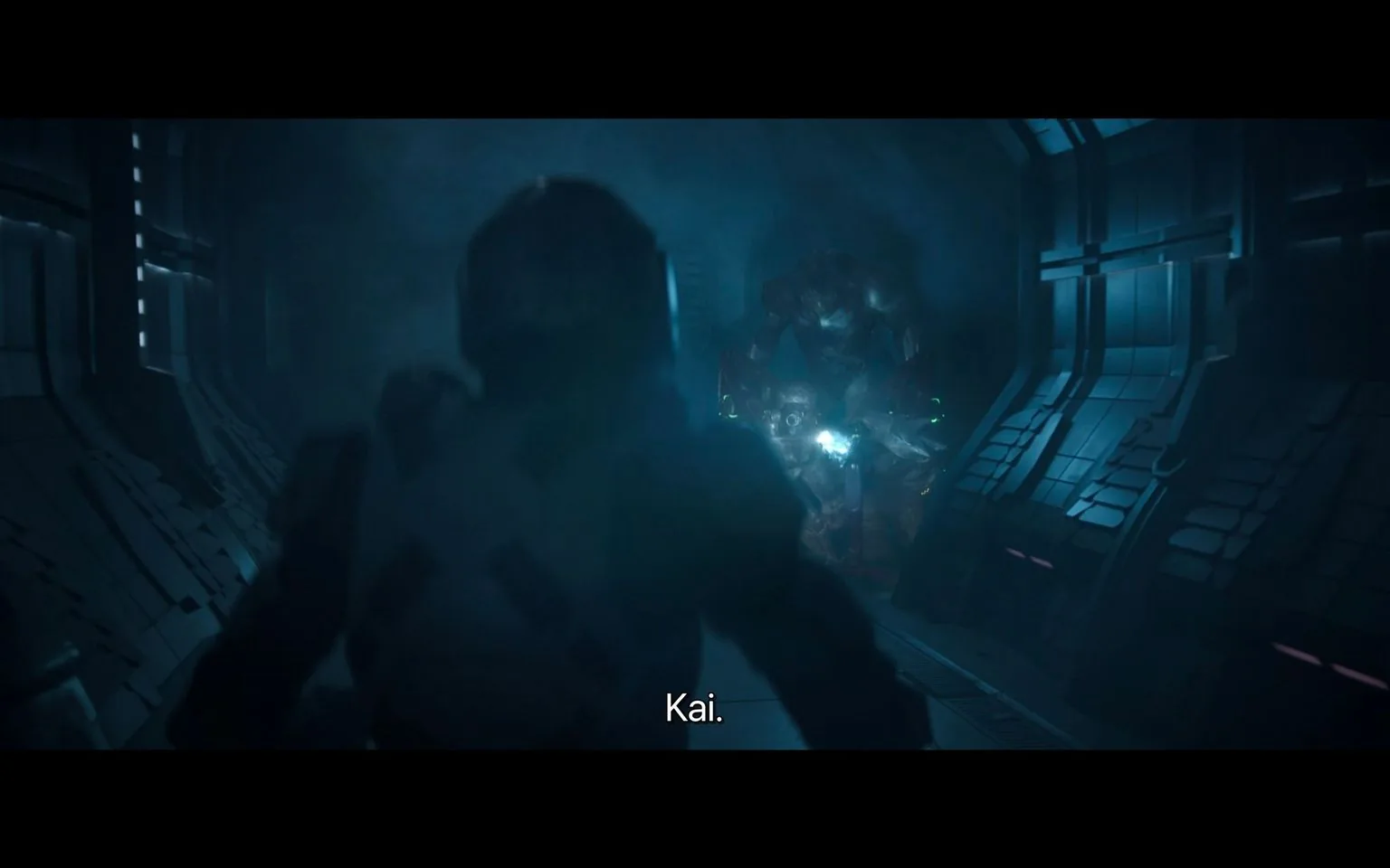 The image is a screenshot from the show "Halo," depicting a tense scene in a dimly lit futuristic corridor. In the foreground, a figure in a suit, presumably of armor, is seen with their back to the viewer, facing a large, imposing creature or figure in the distance. The corridor is metallic, with intricate paneling and a low, moody blue light creating a sense of suspense and danger. The figure in the background appears to be of significant size, adding to the tension of the moment. The environment suggests a high-tech setting, typical of a sci-fi narrative. The subtitle “Kai.” is visible at the bottom of the image, likely indicating a character named Kai is part of the scene. The overall atmosphere is dark and foreboding, contributing to the dramatic tension in the scene.