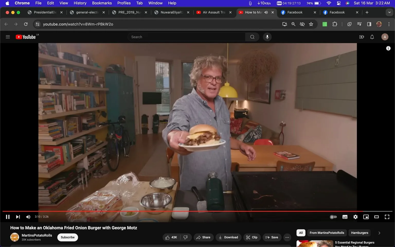 In this YouTube video screenshot, George Motz is presenting an Oklahoma Fried Onion Burger on a plate, extending it towards the camera in a welcoming gesture. The setting appears to be a cozy kitchen or living room, with warm lighting adding a casual and inviting atmosphere. George is dressed in a casual denim shirt, and his expression is friendly and engaging, suggesting he's enthusiastic about sharing his cooking process. Behind him, a shelf filled with books and a bicycle can be seen, along with some homey elements like a couch and a yellow pendant lamp. The table in the foreground displays various cooking ingredients and tools, including a packet of Martin's Potato Rolls, metal bowls, and sliced onions, indicating the preparation process of the burger. The video is titled "How to Make an Oklahoma Fried Onion Burger with George Motz," and it is hosted on the YouTube channel "MartinsPotatoRolls."
- Title: "How to Make an Oklahoma Fried Onion Burger with George Motz"
- Channel / profile: "MartinsPotatoRolls"
- Site / app: "YouTube"